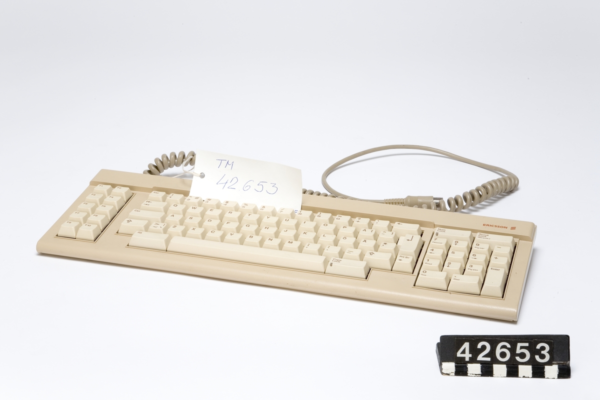 Tangentbord till persondator av beige plast. QWERTY-layout med svenska tecken. Funktionstangeter och numerisk del. Spiralsladd med DIN-kontakt för anslutning till dator.