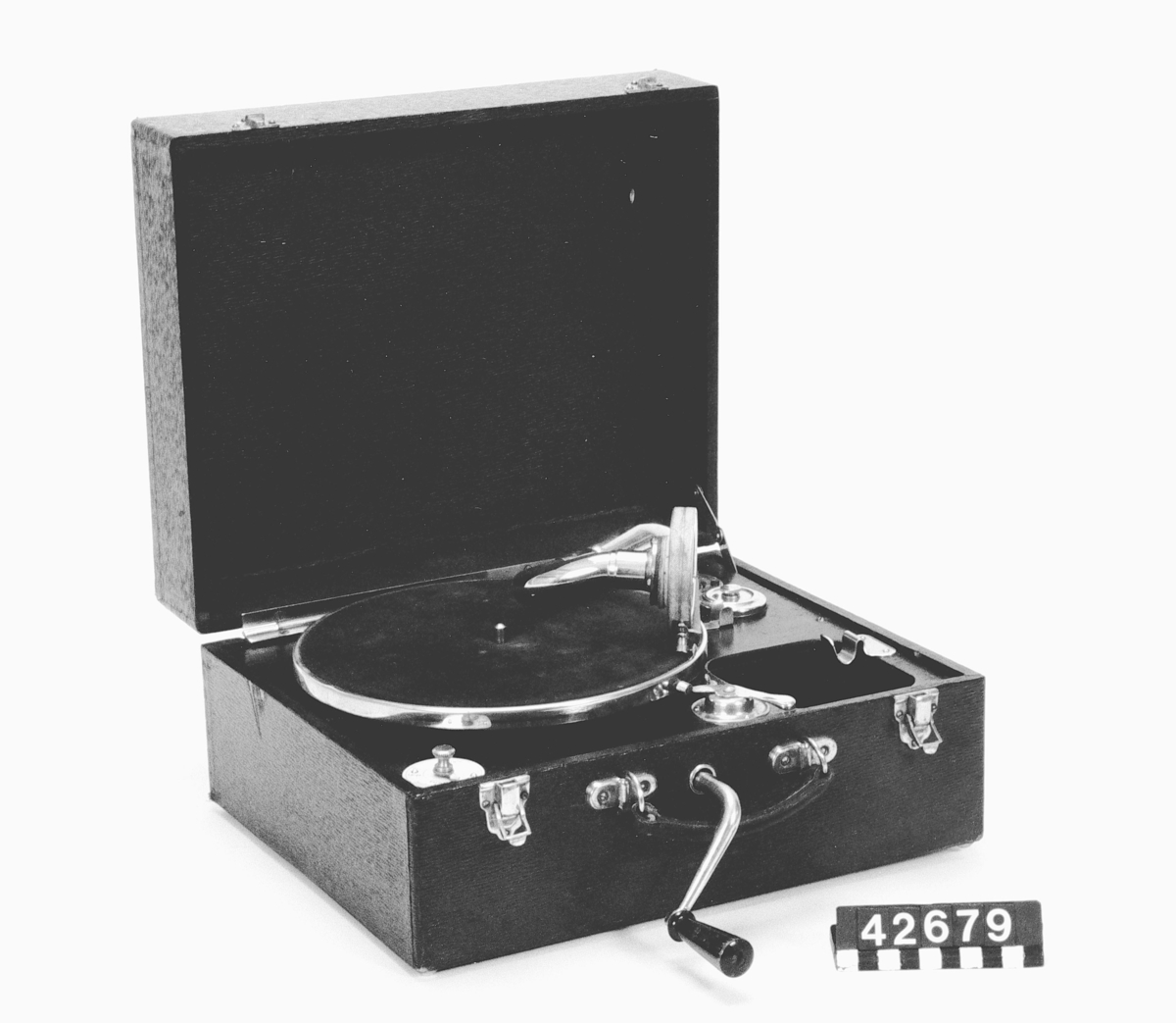 Grammofon av trä och metall, med fjäderverk. Klädd med svart pegamoid. Omärkt ljuddosa.
Tillbehör: Grammofonnål.
