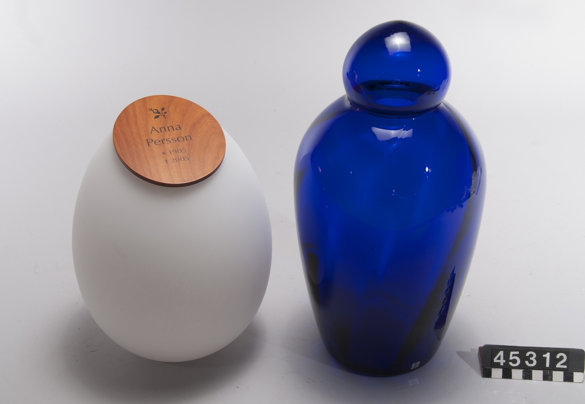 Nedbrytningsbar askurna av patenterat glasmaterial som vittrar sönder efter gravsättning. Då urnan med aska utsätts för markens fukt startar en process som bryter ner glasets grundstruktur. Efter 15 år har glaset omvandlats till grus.  Två urnor - en blå med lock av glas modell Andante samt en matt vit modell Dolce med lock av trä märkt "Anna Persson *1905 +2005", B-37.  Båda märkta "Mats Jonasson, Målerås".