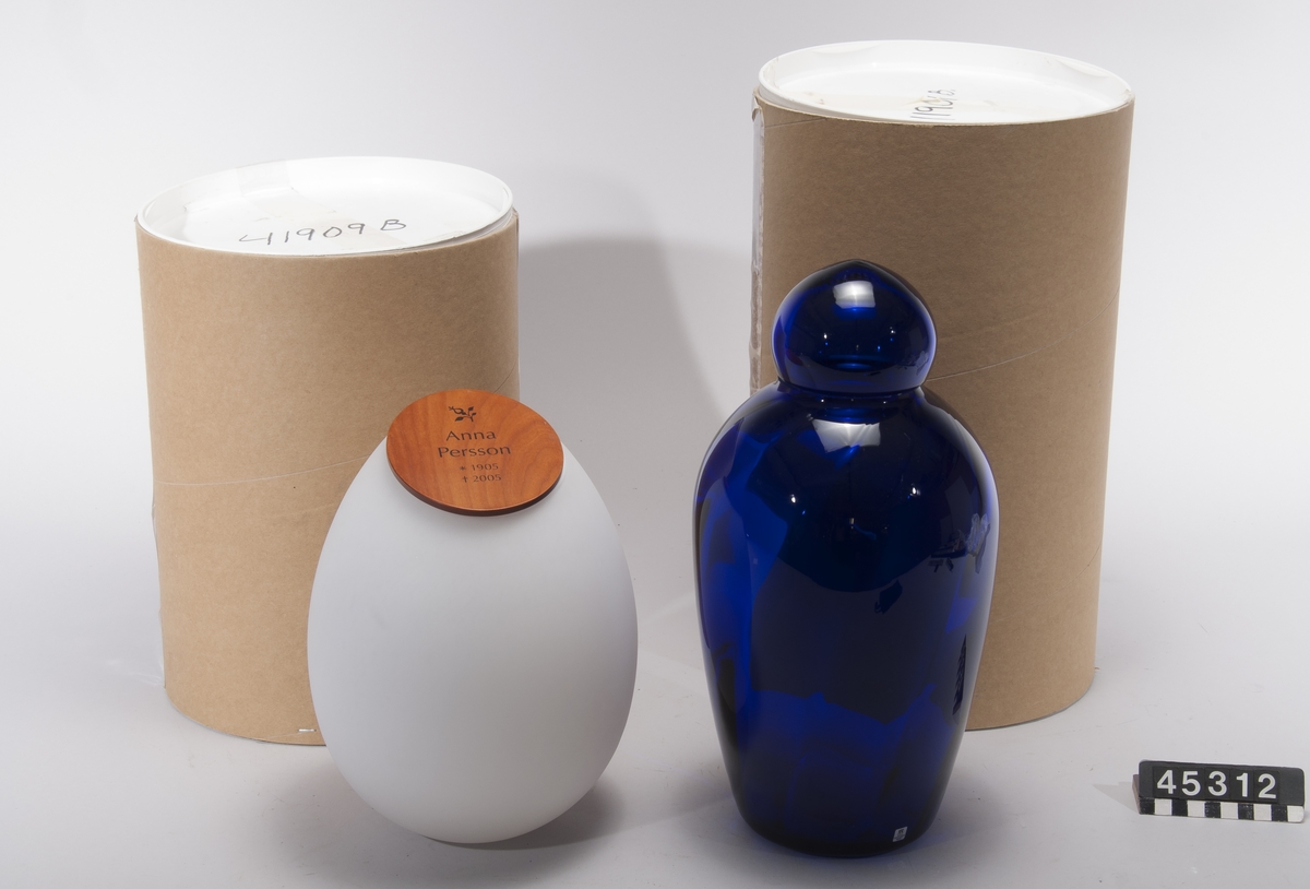 Nedbrytningsbar askurna av patenterat glasmaterial som vittrar sönder efter gravsättning. Då urnan med aska utsätts för markens fukt startar en process som bryter ner glasets grundstruktur. Efter 15 år har glaset omvandlats till grus.  Två urnor - en blå med lock av glas modell Andante samt en matt vit modell Dolce med lock av trä märkt "Anna Persson *1905 +2005", B-37.  Båda märkta "Mats Jonasson, Målerås".