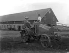 En man och en pojke på en traktor.
Ekonomibyggnader i bakgrunden.