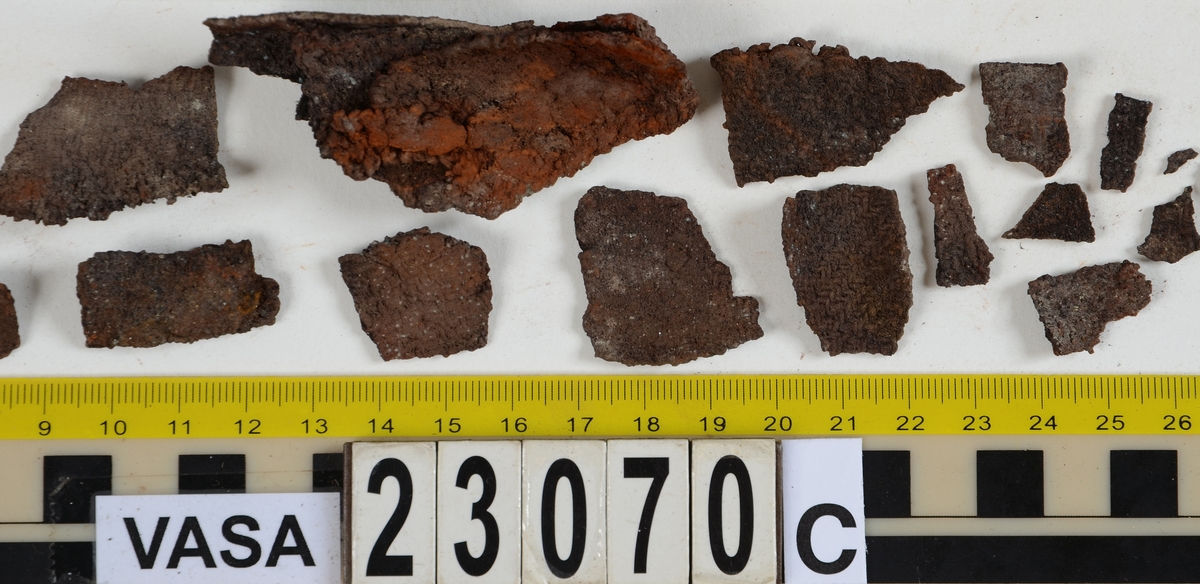 Sko.
Inuti skorna hittades även fragment från olika textilier som ursprungligen varit ett par strumpor. Idag sitter ett stickat fragment av ull fast i vänsterskon (23070) med avigsidan uppåt. I fyndet finns även en mängd lösa stickade fragment som idag är mycket korroderade och vissa helt mineraliserade av järn vilket har gjort textilen stenhård och därmed oerhört skör (23070a-c). Utifrån minskningar som några små fragment har så har det varit möjligt att rekonstruera strumpans tå. Genom att pussla ihop fragmenten som ursprungligen har suttit ihop har det på så vis gått att avgöra hur strumpans tå har varit konstruerad. Då textilen är så fragmentarisk är det inte möjligt att avgöra hur resten av strumpan har sett ut eller varit konstruerad. Däremot så finns det fragment av ett kypertvävt tyg av ull som på vissa ställen ligger dubbelt med det stickade 23070c). Ett av dessa fragment har stelnat i formen så som den suttit på fotens främre del inuti skon. Detta fragment täcker delvis det stickade tyget och har dessutom spår av kaststygn längs en kant. Så som det vävda tyget är placerat på det stickade tyget pekar på att man använt det som lagningslappar till sina stickade strumpor. 
I fyndet finns det även korroderade järnklumpar, ett tuskaftsvävt tyg av ull och rester av ett grovt bastfibergarn och två mindre läderfragment (23070d-f). Det är möjligt att även dessa har tillhört strumpan och/eller skon.

För mer detaljerad information från textildokumentationen (år 2022) se länkade filer.

Skodokumentation 2022:
Ovanläder. Tillverkad i getskinn av god kvalitet. Hör ihop med fyndnummer 23069 (Yngves skor). Tillverkades på en rak läst och bars på vänster fot. Inga dekorationer Det finns stygnhål som visar att det funnits invändiga förstärkningar (men förstärkningslappen saknas). Det finns delaminering men inga lagningar.
För mer information, se Länkade filer: Uppritning

Text in english:
This is the upper of a shoe of more “extreme fashion”, made of goatskin leather of good quality, probably made on a straight last and worn of the left foot, with no decorative features. The upper couples with 23069, together composing Yngve’s shoes. There are internal reinforcements missing from the upper and delamination, there are no repairs.
For more information, go to Länkade filer: Uppritning