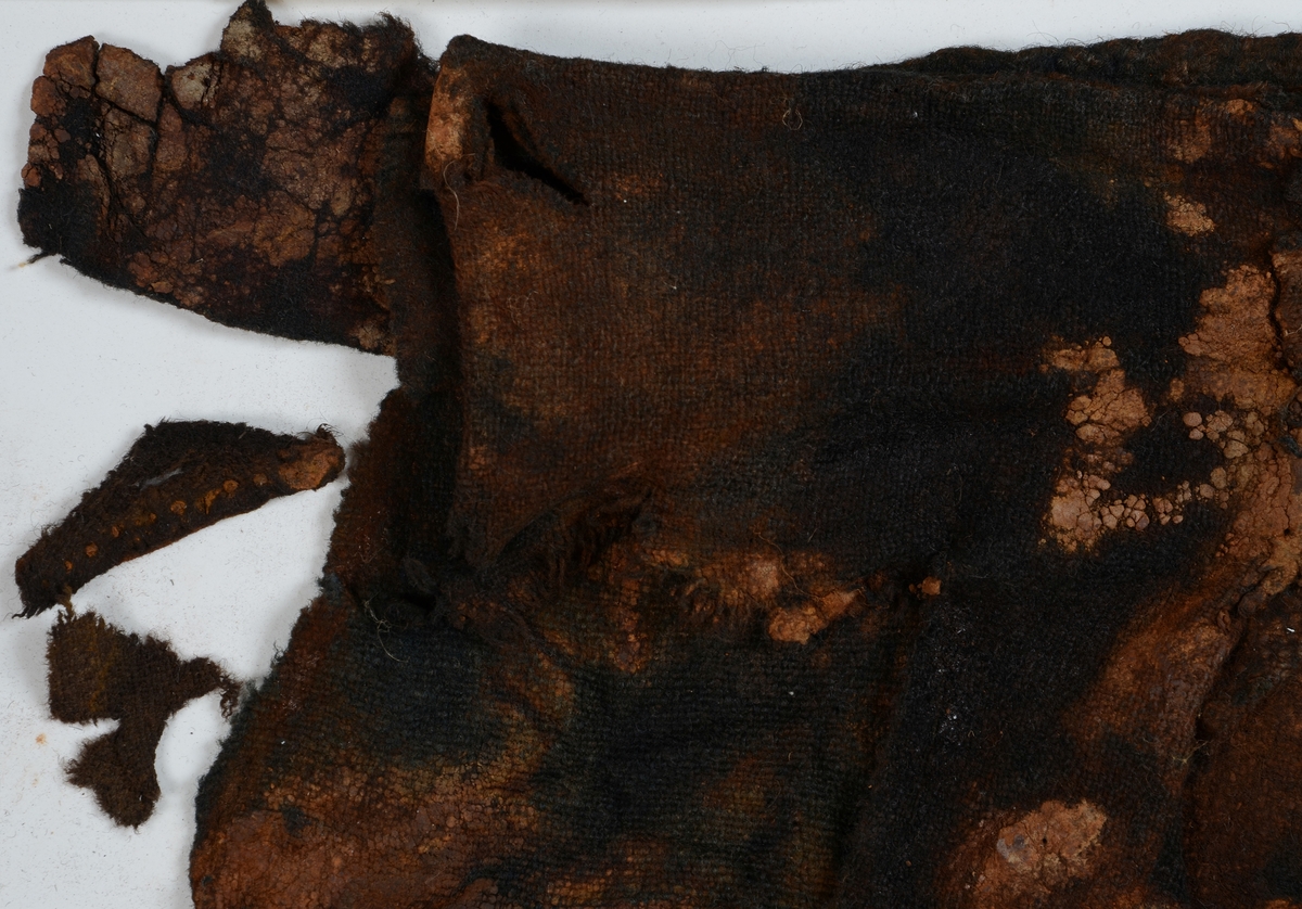 Textil. 13 tygfragment uppdelade på fyndnummer 03466a-d.
Fnr 03466a består av 10 fragment av ull vävda i tuskaft varav det största är ihopsytt av flera delar. Har varit ett par byxor med lucka fram. Originalfärgen kan ha varit blå då rester av blå färg syns på insidan av två av bitarna på de delar där sömmen varit och tyget originalyta bevarats bättre. På en av dessa bitar som tolkats som en del av en ficka på byxans vänstra sida finns rester av en tvåtrådig, s-tvinnad, z-spunnen tråd i sömmen. Byxans tyg har varit mer valkad på insidan än på utsidan. På det största fragmentet framsida syns två knapphål i linningen, och rester efter ytterligare två hål. Det kan därför konstateras att det ursprungligen har varit två knapphål på var sida på linningens mittdel där sidodelarna av linningen knäppts eller knutits fast. Vid ena knapphålet syns något som kan vara resterna av en knapp eller hyska odyl. När byxans delar läggs ihop som de en gång suttit mäter midjebredden ca 47 cm vilket ger ett midjeomfång på ca 94 cm före ihopsnörning. Av det som kvarstår av byxbenen kan konstateras att dagens (år 2015) längd på vänster byxben är 60 cm från linningens övre kant och ner. Höger byxben är i dagsläget endast 43 cm långt. 
På fnr 03466b syns rester av söm i en av kanterna. Fragmentet är av ull och vävt i 2/2-kypert. Lappen har en söm i en cirkel mitt på tyget som troligen är resterna efter den söm som sytt fast lappen runt ett hål på byxan. Kan ha varit en lagningslapp på byxans högra knä som uppvisar motsvarande söm kring ett cirkelformat hål. 
Fnr 03466c är ett fragment av ull vävt i lite finare tuskaftsväv än fnr 03466a. Fragmentet har hål efter sömmar i kanterna samt delar av en ljus tråd i en av kanterna. Blå färg syns där tyget varit skyddat där sömmen varit. Fragmentet kan ha varit en innerficka bak på byxans vänstra sida. Det finns en söm i byxans tyg där som överensstämmer med lappens söm.
Alla kanterna på fnr 03466d är invikta och har hål efter söm som om biten varit fastsydd på ett annat tygstycke. Fragmentet är av ull och vävt i samma sorts tuskaftsväv som byxan. Kan ha varit en lagningslapp på byxan över ett hål i byxtyget på den plats där vänster innerficka bak är placerad.