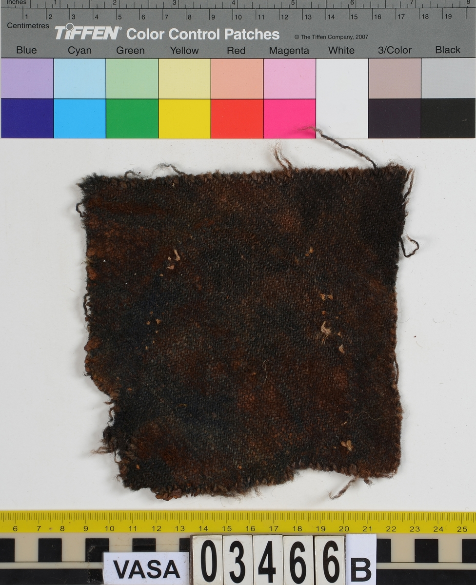 Textil. 13 tygfragment uppdelade på fyndnummer 03466a-d.
Fnr 03466a består av 10 fragment av ull vävda i tuskaft varav det största är ihopsytt av flera delar. Har varit ett par byxor med lucka fram. Originalfärgen kan ha varit blå då rester av blå färg syns på insidan av två av bitarna på de delar där sömmen varit och tyget originalyta bevarats bättre. På en av dessa bitar som tolkats som en del av en ficka på byxans vänstra sida finns rester av en tvåtrådig, s-tvinnad, z-spunnen tråd i sömmen. Byxans tyg har varit mer valkad på insidan än på utsidan. På det största fragmentet framsida syns två knapphål i linningen, och rester efter ytterligare två hål. Det kan därför konstateras att det ursprungligen har varit två knapphål på var sida på linningens mittdel där sidodelarna av linningen knäppts eller knutits fast. Vid ena knapphålet syns något som kan vara resterna av en knapp eller hyska odyl. När byxans delar läggs ihop som de en gång suttit mäter midjebredden ca 47 cm vilket ger ett midjeomfång på ca 94 cm före ihopsnörning. Av det som kvarstår av byxbenen kan konstateras att dagens (år 2015) längd på vänster byxben är 60 cm från linningens övre kant och ner. Höger byxben är i dagsläget endast 43 cm långt. 
På fnr 03466b syns rester av söm i en av kanterna. Fragmentet är av ull och vävt i 2/2-kypert. Lappen har en söm i en cirkel mitt på tyget som troligen är resterna efter den söm som sytt fast lappen runt ett hål på byxan. Kan ha varit en lagningslapp på byxans högra knä som uppvisar motsvarande söm kring ett cirkelformat hål. 
Fnr 03466c är ett fragment av ull vävt i lite finare tuskaftsväv än fnr 03466a. Fragmentet har hål efter sömmar i kanterna samt delar av en ljus tråd i en av kanterna. Blå färg syns där tyget varit skyddat där sömmen varit. Fragmentet kan ha varit en innerficka bak på byxans vänstra sida. Det finns en söm i byxans tyg där som överensstämmer med lappens söm.
Alla kanterna på fnr 03466d är invikta och har hål efter söm som om biten varit fastsydd på ett annat tygstycke. Fragmentet är av ull och vävt i samma sorts tuskaftsväv som byxan. Kan ha varit en lagningslapp på byxan över ett hål i byxtyget på den plats där vänster innerficka bak är placerad.