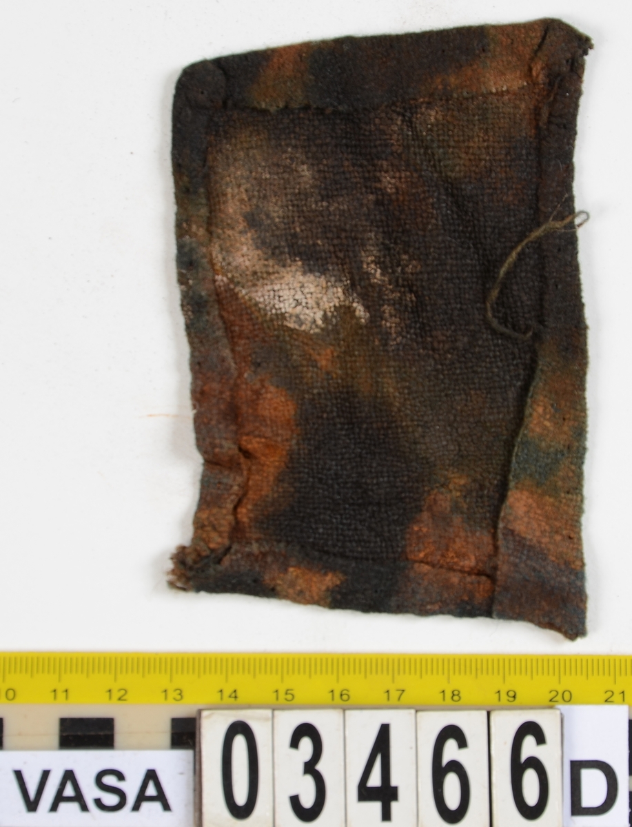 Textil. 13 tygfragment uppdelade på fyndnummer 03466a-d.
Fnr 03466a består av 10 fragment av ull vävda i tuskaft varav det största är ihopsytt av flera delar. Har varit ett par byxor med lucka fram. Originalfärgen kan ha varit blå då rester av blå färg syns på insidan av två av bitarna på de delar där sömmen varit och tyget originalyta bevarats bättre. På en av dessa bitar som tolkats som en del av en ficka på byxans vänstra sida finns rester av en tvåtrådig, s-tvinnad, z-spunnen tråd i sömmen. Byxans tyg har varit mer valkad på insidan än på utsidan. På det största fragmentet framsida syns två knapphål i linningen, och rester efter ytterligare två hål. Det kan därför konstateras att det ursprungligen har varit två knapphål på var sida på linningens mittdel där sidodelarna av linningen knäppts eller knutits fast. Vid ena knapphålet syns något som kan vara resterna av en knapp eller hyska odyl. När byxans delar läggs ihop som de en gång suttit mäter midjebredden ca 47 cm vilket ger ett midjeomfång på ca 94 cm före ihopsnörning. Av det som kvarstår av byxbenen kan konstateras att dagens (år 2015) längd på vänster byxben är 60 cm från linningens övre kant och ner. Höger byxben är i dagsläget endast 43 cm långt. 
På fnr 03466b syns rester av söm i en av kanterna. Fragmentet är av ull och vävt i 2/2-kypert. Lappen har en söm i en cirkel mitt på tyget som troligen är resterna efter den söm som sytt fast lappen runt ett hål på byxan. Kan ha varit en lagningslapp på byxans högra knä som uppvisar motsvarande söm kring ett cirkelformat hål. 
Fnr 03466c är ett fragment av ull vävt i lite finare tuskaftsväv än fnr 03466a. Fragmentet har hål efter sömmar i kanterna samt delar av en ljus tråd i en av kanterna. Blå färg syns där tyget varit skyddat där sömmen varit. Fragmentet kan ha varit en innerficka bak på byxans vänstra sida. Det finns en söm i byxans tyg där som överensstämmer med lappens söm.
Alla kanterna på fnr 03466d är invikta och har hål efter söm som om biten varit fastsydd på ett annat tygstycke. Fragmentet är av ull och vävt i samma sorts tuskaftsväv som byxan. Kan ha varit en lagningslapp på byxan över ett hål i byxtyget på den plats där vänster innerficka bak är placerad.