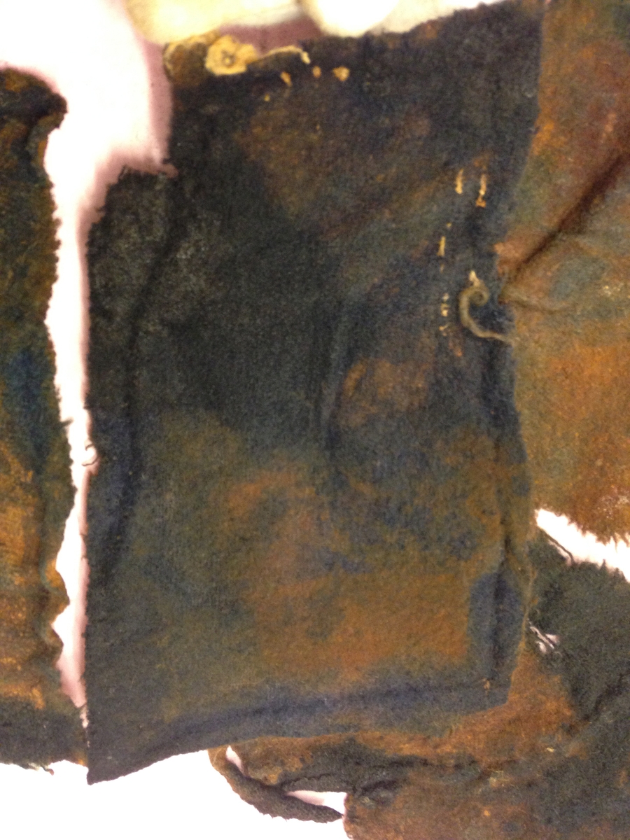 Textil. 13 tygfragment uppdelade på fyndnummer 03466a-d.
Fnr 03466a består av 10 fragment av ull vävda i tuskaft varav det största är ihopsytt av flera delar. Har varit ett par byxor med lucka fram. Originalfärgen kan ha varit blå då rester av blå färg syns på insidan av två av bitarna på de delar där sömmen varit och tyget originalyta bevarats bättre. På en av dessa bitar som tolkats som en del av en ficka på byxans vänstra sida finns rester av en tvåtrådig, s-tvinnad, z-spunnen tråd i sömmen. Byxans tyg har varit mer valkad på insidan än på utsidan. På det största fragmentet framsida syns två knapphål i linningen, och rester efter ytterligare två hål. Det kan därför konstateras att det ursprungligen har varit två knapphål på var sida på linningens mittdel där sidodelarna av linningen knäppts eller knutits fast. Vid ena knapphålet syns något som kan vara resterna av en knapp eller hyska odyl. När byxans delar läggs ihop som de en gång suttit mäter midjebredden ca 47 cm vilket ger ett midjeomfång på ca 94 cm före ihopsnörning. Av det som kvarstår av byxbenen kan konstateras att dagens (år 2015) längd på vänster byxben är 60 cm från linningens övre kant och ner. Höger byxben är i dagsläget endast 43 cm långt. 
På fnr 03466b syns rester av söm i en av kanterna. Fragmentet är av ull och vävt i 2/2-kypert. Lappen har en söm i en cirkel mitt på tyget som troligen är resterna efter den söm som sytt fast lappen runt ett hål på byxan. Kan ha varit en lagningslapp på byxans högra knä som uppvisar motsvarande söm kring ett cirkelformat hål. 
Fnr 03466c är ett fragment av ull vävt i lite finare tuskaftsväv än fnr 03466a. Fragmentet har hål efter sömmar i kanterna samt delar av en ljus tråd i en av kanterna. Blå färg syns där tyget varit skyddat där sömmen varit. Fragmentet kan ha varit en innerficka bak på byxans vänstra sida. Det finns en söm i byxans tyg där som överensstämmer med lappens söm.
Alla kanterna på fnr 03466d är invikta och har hål efter söm som om biten varit fastsydd på ett annat tygstycke. Fragmentet är av ull och vävt i samma sorts tuskaftsväv som byxan. Kan ha varit en lagningslapp på byxan över ett hål i byxtyget på den plats där vänster innerficka bak är placerad.