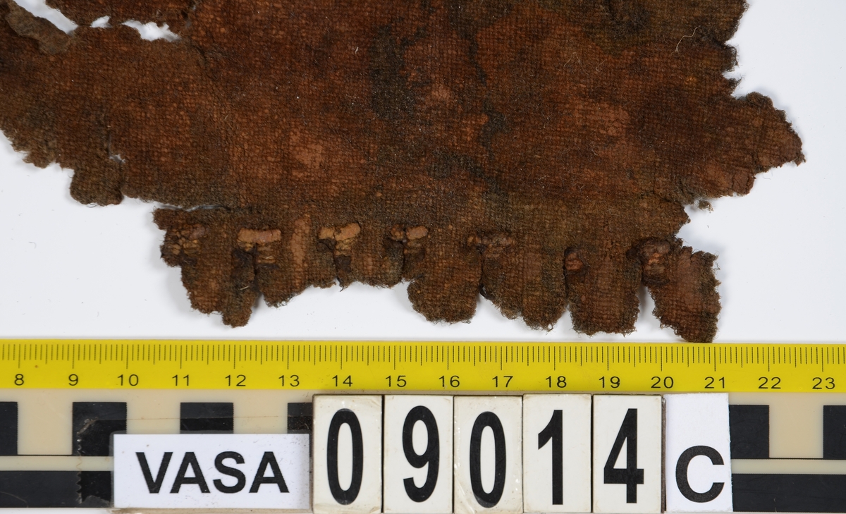Textildokumentation 2018
Textil, fyndnumret består av mer än 50 st fragment uppdelade på 09014a-h.
Kontextgrupp ÖB20 (övriga fyndnummer i gruppen: 07732, 07762, 09228)
Relaterade fynd: Kontextgrupp ÖB17 och ÖB 18.
.
Fnr 09014a Typnr ÖB20.3:1
Sammanfattning - Ett textilfragment av ull vävt i tuskaft. Färgen är nu mörkbrun, fibrerna varierar i färg vilket tyder på att det är naturfärgad ull. Fibrerna är i vissa partier skadade på ytan och ser därför vita ut. Väven är tunn, jämn och fin och har inga spår av ytbehandling. Fragmentet har tre tillskurna kanter, två av dessa har spår av tre olika sömmar (söm 1: kvicksöm/förstygn). Formen tyder på att det är ett skört till en tröja. Andra delar: Jämför med 07726. 
.
Fnr 09014a Typnr ÖB20.4:1
Sammanfattning - Ett textilfragment av ull vävt i tuskaft. Färgen är nu mörkbrun, originalfärg okänd. I mikroskop ser tyget blåsvart ut. Väven är mycket jämn och slät. I sömsmånen på sida A finns bevarad lugg, på sida B är luggen så välbevarad att bindningen inte syns. Fragmentet har två tillskurna kanter med hål och avtryck av sömmar, även trådrester finns (söm 1: kaststygn - fälld kant, söm 2: ev förstygn - sammanfogande, söm 3: kaststygn - fälld kant). På sida B finns blåfärgad ull och silkesfibrer. 
.
Fnr 09014a Typnr ÖB20.5:1
Sammanfattning - Ett textilfragment av ull vävt i tuskaft. Väven är jämn och tät. En av kanterna är troligen tillskuren, inga sömrester finns. Tyget har inga spår av ytbehandling. På ytan finns en del bastfibrer. Relaterade fynd: Vävens struktur liknar ÖB20.4:1 men tygerna har ändå olika karaktär och detta fragment saknar lugg. 
.
Fnr 09014a Typnr ÖB20.6:1
Sammanfattning - Ett textilfragment av ull vävt i tuskaft. Färgen är nu brun, originalfärg är okänd men möjligen naturfärgad, fibrerna är mycket släta och varierar i färg från brun till nästan svart. Fragmentet är litet och mycket skadat, endast några få bindepunkter återstår vilket försvårar mätningar. 
.
Fnr 09014a Typnr ÖB20.7:1
Sammanfattning - Ett textilfragment av  ull vävt i tuskaft. Färgen är svartbrun, originalfärg okänd men möjligen naturfärgad då fibrernas färg varierar. Inga tydliga spår av ytbehandling. Väven är jämn men garnet är hårt spunnet vilket ger tyget en kypertkaraktär (falsk kypert). Fragmentet har inga tillskurna kanter eller sömmar. På ytan finns enstaka vita silkesfibrer.
.
Fnr 09014a Typnr ÖB20.8:1
Sammanfattning - Ett textilfragment av ull. Fragmentet är mycket litet och dåligt bevarat så bindningen är svår att avgöra, troligen 2/2 kypert. Färgen är svart, möjligen naturfärgad ull då fibrerna varierar något i färg. Tyget har ingen synlig ytbehandling. Garnet är hårt spunnet vilket ger tyget en speciell karaktär. 
.
Fnr 09014b Typnr ÖB17.1:8-11
Sammanfattning - Fyra textilfragment av ull vävt i tuskaft. Färgen är mörkbrun, originalfärg okänd men den bruna tonen slår mot röd. Tre av fragmenten har tillskurna kanter och spår av sömmar. Andra delar, som troligen kommer från samma tröja, är fnr 03261a och d, 09014h samt 09228a. 
Fragment ÖB17.1:9 - Fragmentet har två tillskurna kanter varav den ena har spår av en söm. Fragmentet är troligen sidostycket till en tröja. Den tillskurna kanten har då varit monterad mot tröjans bakstycke och bör ha hört samman med fyndnummer 03261d, vilket är ett bakstycke till en tröja.
Fragment ÖB17.1:10 - Fragmentet är mycket skadat men är troligen av typen ÖB17.1. Formen är densamma som för del av fragment ÖB17.1:9 och kan därför vara en dubblering av detsamma, alternativt det motstående sidostycket.
.
Fnr 09014c Typnr ÖB20.9:1-19
Sammanfattning - Nitton textilfragment av ull vävt i tuskaft. Färgen är brun, originalfärg okänd. Sida B har en kort tät lugg som döljer bindningen. På sida A finns ingen lugg och bindningen är synlig. Fjorton av fragmenten har tillskurna kanter och sömmar. Tre av fragmenten har sydda knapphål. Flera fragment är dräktdelar, till exempel finns en fragmentarisk krage. På ytan på flertalet fragment finns fibrer i flera klara färger, till exempel rosa, röd och blå. Vita fibrer finns i stor mängd, kan vara recenta.

Fragment ÖB20.9:1 - Fragmentet har flera tillskurna kanter med avtryck och hål efter olika typer av sömmar (söm 1: ev. sammanfogande, söm 2: kaststygn - fåll, söm 3: kaststygn - sammanfogande, söm 4: kaststygn - sammanfogande, söm 5: osäkert, söm 6: osäkert), trådrester finns. Kanterna och sömmarnas karaktär gör att sida A bedöms vara plaggets rätsida/utsida. Några hål som ej är stygnhål men kan vara original finns. I fragmentet sitter fast två stycken silkestrådar (söm 7: silkestråd med knut, fäst i tyget, söm 8:  silkestråd med ögla, fäst i tyget). Det är samma typ av tråd som knapphålen på fragment ÖB20.9:2 är sydda med. På sida A finns ett område med extra mycket korrosion och även en långsmal vitaktig avlagring.

Fragment ÖB20.9:2 - Fragmentet har inga bevarade tillskurna kanter, dock finns en någorlunda välbevarad rad med knapphål. Samtliga knapphål är trasiga i ena sidan så deras ursprungliga storlek går ej att avgöra. Knapphålen är sydda med knapphålsstygn i silkestråd och sitter med 13-15 mm mellanrum (c/c mått). Det tvärställda tränsen består av ca 10 stygn och syns ej på sida B, tränsens knutar sitter vända från knapphålet. På sida B anas rester av bastfiber under knapphålsstygnen, troligen från en dubblering av tyget eller ett foder. 

Fragment ÖB20.9:3 - Fragmentet har fyra stycken knapphål, varav ett är helt. Knapphålen är skurna på skrådden. Eventuellt finns spår av en söm längs en del av kanten. Knapphålsstygn och trådrester är mycket fragmentariska.

Fragment ÖB20.9:4 - Fragmentet har tre stycken knapphål.  Knapphålen är sydda med knapphålsstygn i silkestråd. Knapphålens längd är 19-20 mm och de sitter med 18-22 mm mellanrum (c/c mått). Eventuellt är fragmentets ena kant en sömsmån, kan ha varit monterad mellan andra tyglager (kanten har avvikande färg). Under knapphålsstygnen på sida B finns rester av annat material, eventuellt bastfiber. Bastfibrer finns också utspridda över ytan på sida B, kan vara rester av en dubblering till tyget eller ett foder, se fragment 9:2. På sida A finns avlagringar och korrosion. 

Fragment ÖB20.9:5 - Fragmentet har bristningar i tyget som ser ut som knapphål, dock finns inga rester av stygnhål eller tråd. Fragmentets ena kant är troligen en sömsmån, kan ha varit monterad mellan andra tyglager (kanten har avvikande färg). Enstaka spår av söm och tråd finns längs kanten (söm 1), dessa är mycket otydliga. På fragmentets andra (mycket trasiga) kant finns ett mycket tydligt stygnhål. Fragment 9:4 och 9:5 ser ut att kunna höra ihop, dock går trådsystemen åt olika håll så det är inte fallet.

Fragment 9:6-9 - Till det stora fragmentet 9:6 hör det lilla lösa fragmentet 9:7. I fragment 9:6 sitter två fragment fastsydda, 9:8 och 9:9. De sammansydda fragmenten har tre tillskurna kanter och spår av minst fem sömmar (söm 1: kaststygn, söm 2: förstygn el. liknande - sammanfogande, söm 3: kaststygn, söm 4: sammanfogande, fäster ihop 9:6 med 9:8 och 9:9, söm 5: två stygnhål), trådrester finns. Det är troligt att 9:8 och 9:9 är delar av samma tillskurna bit, och att den utgjort en dubblering till 9:6 (inklusive 9:7). Den tillskurna formen ser ut att vara en krage. Fragmentet skulle då vara en halv krage, bevarad från framkant till nästan mitt bak. På sida A finns en beläggning på delar av ytan. På sida B finns en mängd fibrer i klara färger: blå, vit, rosa, tegelröd, orange, karminröd, grön, mörkgul och grå. fibrerna ligger ytligt med nästan samtliga sitter delvis fast i tygets lugg eller har en eller båda ändar nere i vävens bindning. Fibrerna bör därför vara ursprungliga. Sida A har en mindre mängd av nämnda fibrer, mest förekommande på ytan av fragment 9:8 och 9:9.

Fragment ÖB20.9:10 - Fragmentet har två tillskurna kanter. Dessa är invikta mot sida B som en 6-7 mm bred fåll som är fastsydd med kaststygn, trådrester finns (söm 1 och 2). Fragmentet kan vara ett skört till en tröja. På båda sidors yta finns fibrer i klara färger: grå, ljusblå, orange, orangebrun, vitgul, blå svart, vit, röd, korallröd och rosa.

Fragment ÖB20.9:11 - Fragmentet har tre tillskurna kanter med sömmar. Två av kanterna är invikta och fastsydda mot sida B, som fållar (söm1: kaststygn - fåll, söm 2: kaststygn - fåll). Stygnen går ej igenom till sida A men sömmen syns som bucklor i fragmentets yta. Söm 3 längs den tredje kanten har troligen varit en sammanfogande söm sydd med kaststygn. Stygn och trådrester finns bevarade från samtliga sömmar. Under fållen vid söm 1 finns ett litet fragment av en annan textiltyp. Troligen ursprungligen en dubblering eller liknande. Fragmentet är ca 10x5 mm stort och har fått typnummer ÖB20.16:1. På ytan på sida B finns enstaka bastfibrer.

Fragment ÖB20.9:12 - Fragmentet har två tillskurna kanter med sömmar (söm 1: ev kaststygn, söm 2: osäker). Vid söm 2 är sömsmånen vikt mot sida B. Vid söm 1 är kanten inte vikt. Fragmentet har samma form som ÖB20.9:6, "kragen".

Fragment ÖB20.9:13 - Fragmentet har två tillskurna kanter med sömmar (söm1: sammanfogande, söm 2: osäkert). De två övriga kanterna är eventuellt också de tillskurna men spår av eventuella sömmar är för otydliga. Enstaka fibrer med klara färger (rosa, blå, vit) finns på båda sidor, fler på sida B. Vid söm 2 finns på ett ställe en fiberknippa som kommer upp ur vävens bindning, kan vara rester av en tråd och är eventuellt halsplat silke. 

Fragment ÖB20.9:14 - Fragmentet har en tillskuren kant med söm (söm1: ev sammanfogande). I stygnhålen finns rester av tråd.
.
Fnr 09014c Typnr ÖB20.10:1
Sammanfattning - Ett textilfragment av ull vävt i tuskaft. Färgen är brun, originalfärg okänd. Sida B har en kort och tät lugg, bindningen syns bara på sida A. Längs ena kanten finns tre stycken tydliga hål. Hålen är gjorda med ett verktyg som skär av fibrerna snarare än tränger undan dem.
.
Fnr 09014c Typnr ÖB20.16:1
Sammanfattning - Fragmentet är mycket litet och sitter inuti den invikta fållen på fragment 09014c ÖB20.9:11, det sticker bara fram någon millimeter. Fragmentet är mycket skört. Bevarandegrad och storlek gör mätningar svåra. Det är olika garn i trs 1 och trs 2. Sida B är täckt av en lugg, på sida A syns bindningen, om än fragmentariskt.
.
Fnr 09014d Typnr ÖB18.16:2-8
Ej dokumenterad
.
Fnr 09014d Typnr ÖB20.11:1-2
Ej dokumenterad
.
Fnr 09014e Typnr ÖB20.12:1-4
Sammanfattning - Fyra textilfragment av ull vävt i tuskaft. Färgen är brun med inslag av täckhårsfibrer i en mörkare brun färg, originalfärg okänd. Fragmenten är mycket fragmentariska med trs 1 bättre bevarat än trs 2. Enbart på en del av fragment ÖB20.12:1 finns trs 2 samt bindningen bevarad. Utspridda över tygets yta finns grova bastfibrer samt korrosionsfläckar. 
.
Fnr 09014f Typnr ÖB20.13:1
Sammanfattning - Ett textilfragment av ull vävt tuskaft. Färgen är mörkbrun, fibrerna varierar mellan ljus och mörk brun. Originalfärg okänd, möjligen naturfärgad ull. Garnet är hårt spunnet vilket ger väven en särskild karaktär, sida A har en mer strukturerad ytan än sida B. Fragmentet har inga tillskurna kanter. På sida A finns enstaka röda och gulgröna silkesfibrer, samt bastfibrer. På sida B finns enstaka blå, vita och rosa silkesfibrer.
.
Fnr 09014g Typnr ÖB20.14:1-3
Sammanfattning - Tre textilfragment av ull och bastfiber vävt i tuskaft. Färgen är beige för bastfibern (trs 1) och svartblå för ullen (trs 2). Trs 2 är mycker grövre än trs 1. Trs 1 är nästan helt försvunnet. Två av fragmenten har tillskurna kanter och ett har spår av en sömsmån. På ytan av fragmenten finns en stor mängd bastfiber samt en vit/rosa tuss med silke. På fragment 14:2 finns några intensivt blå ullfibrer.
.
Fnr 09014h Typnr ÖB17.1:12-13
Sammanfattning - Två textilfragment av ull vävt i tuskaft. Färgen är brun, originalfärg okänd. Det större fragmentet är tillskuret och har spår av sömmar. Fragmentet är en ensömsärm med sömmen placerad mitt bak på ärmen. På ytan finns stora rostfläckar. Andra delar: Hör troligen (trots färgskillnad) till skelettet Beatas tröja, se fnr 03261a-d, 09228a, 09014b och h.