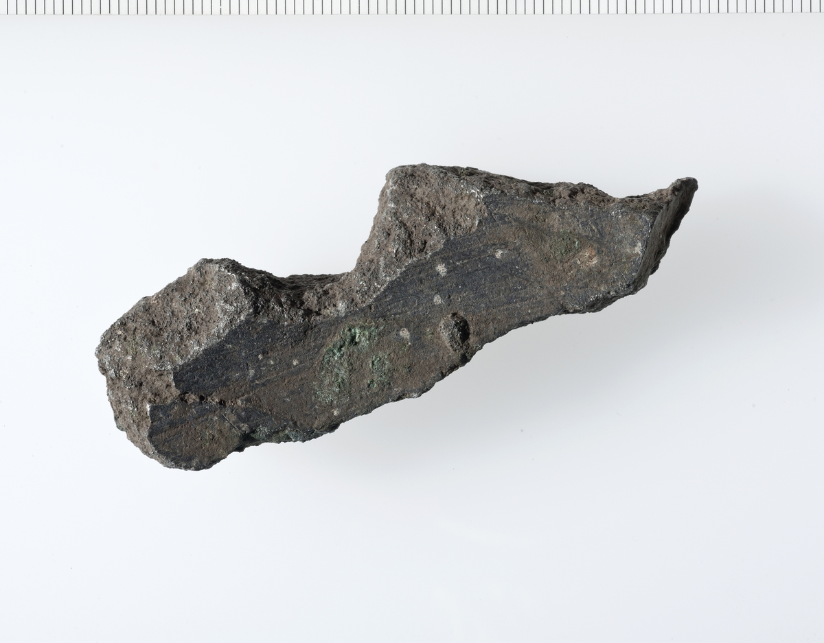 Svakt buet avlangt fragment av støpt materiale. Fragmentets buede sider er glatte, bruddsidene er porøse.  Fragmentet har for tykke vegger til å være fra en gryte, og gir en for stor diameter til å kunne være fra noen kanon. Derfor antatt fragment fra klokke. 