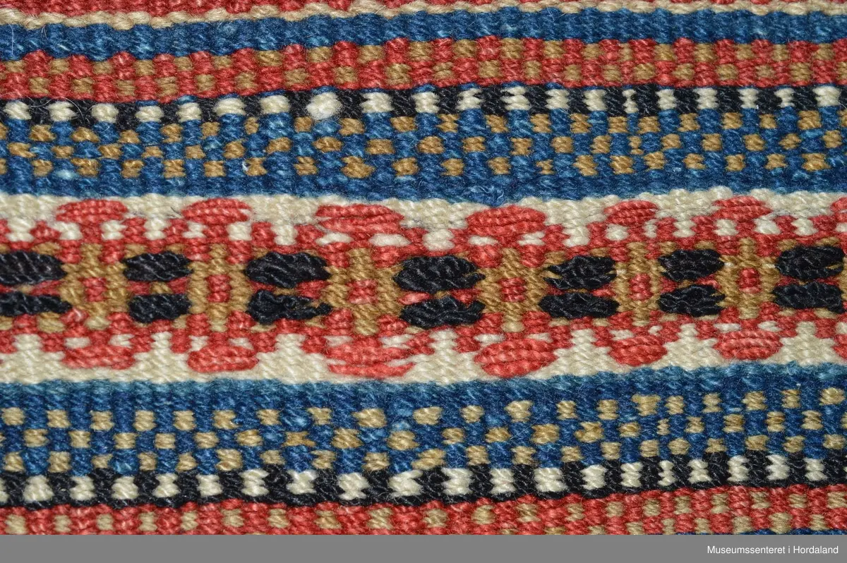 Åkle som er samansett av 8 border i halvsmett og enklare border innimellom.Bukta på renningen ser ein oppe på teppet. Dei første innslaga er delivis bortsletne. Nede på teppet er det kasta over for hand; 1-2 cm lange frynser.