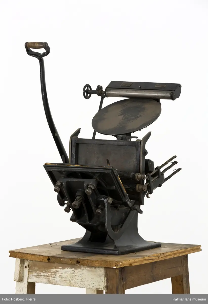 KLM 45377:60. Tryckpress, Trä och metall. Digelpress av svartmålad metall, stommen av gjutjärn. På digelpressens övre del sitter två metallplåtar. På den ena plåten står det Maschinenfabrik A Hogenforst Leipzig. På den andra står det Importerad av Gust. Carlsson&Co Maskinaffär Stockholm. Digelpressen är monterad på ett litet träbord som förmodligen är byggt för pressen. Det är delvis målat i vitt.