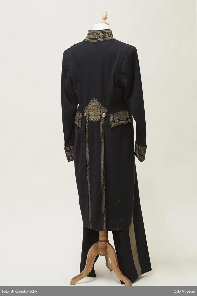 Gallauniform til ambassadør.
A: Jakke-kjole. Jakken er ettersittende foran m. høy krage som lukkes m. hekter. Lang snipp bak. Dekorbånd langs sidene og langs snipp. Dekorerte lommeklaffer og dekor på midten bak. Ermer m. dekorerte mansjettoppslag. Hemper til medaljer. Gullgalloner på buksen.Dekormotiv: Rike blomsterranker m. ekeblad. Lengde foran: 41 cm.
Lengde bak: 114 cm.
B. Bukse m. nesten rette ben. Stikklommer på siden som er skjult under gallongene. Gylf m. knepping. Liten splitt i linning bak. Knapper til seler, doble foran, enkle bak. Dekorert m. gallongbånd i damaskvev. Motiv: Ekebladranker. Lengde:115 cm.
C.Dobbelsnutehatt til ambassadøruniform m. strutsefjær og gullbånd.Stoffet på brem er trukket over et papplikninde materiale. Bremmen er formet opp på sidene slik at hatten former snuter foran og bak. En "Flaggrose" er applikert på høyre side. Tvunnet gullbånd og knapp er festet over. Gulldekor på hver snute. Strutsefjær montert langs hattebrem. Gullbånd m. ekebladmotiv langs hattebrem. Høyde: 13 cm. Lengde: 46 cm. Materiale: Fløyel og papp.God tilstand. Skjør strutsefjær.
D. Hatteeske til dobbelsnutehatt i lakkert lerretstrie/seilduk. Sort. Lukkes m. lærrem på toppen. Esken har hattens form. Vattert, gult bommulsfòr inni.
