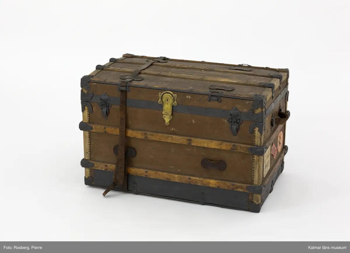 KLM 28581. Koffert, amerikakoffert, trä, väv, metall, läder. Amerikakoffert av trästomme klädd med väv. Trärribbor som är sammanfogade med plåtbeslag finns på alla sidor. Kistan hålls stängd med två bygelbeslag i metallplåt samt ett lås av mässing. Runt kistan har det funnits två läderremmar, endast en av dem finns kvar. På kistans sidor finns bärhandtag av läder. Invändigt är kistan klädd med ett vitt tyg med ränder i gult och blått som bildar rutmönster. En lös insats med tre mindre fack finns i kistans övre del. Den är av trä och klädd med tyg. Överst i kistan finns en löst lock av kartong klädd med samma tyg som resten av kistans insida.