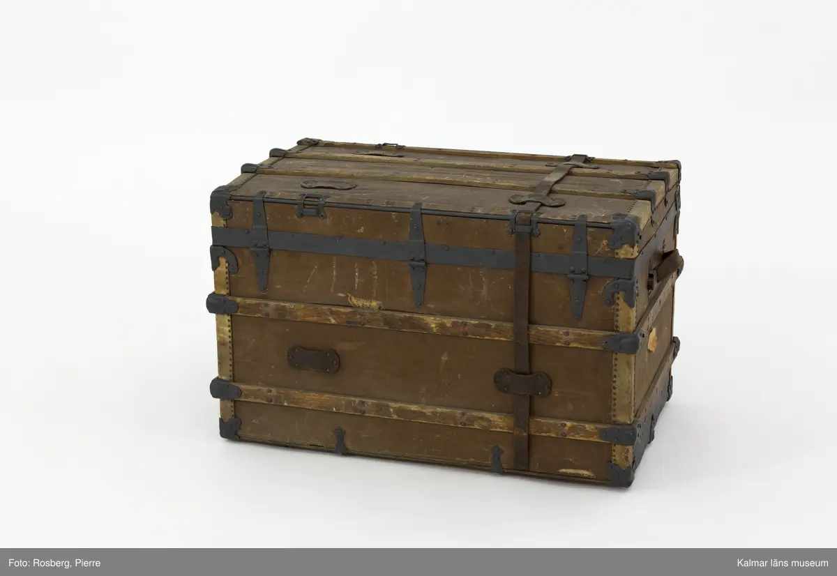 KLM 28581. Koffert, amerikakoffert, trä, väv, metall, läder. Amerikakoffert av trästomme klädd med väv. Trärribbor som är sammanfogade med plåtbeslag finns på alla sidor. Kistan hålls stängd med två bygelbeslag i metallplåt samt ett lås av mässing. Runt kistan har det funnits två läderremmar, endast en av dem finns kvar. På kistans sidor finns bärhandtag av läder. Invändigt är kistan klädd med ett vitt tyg med ränder i gult och blått som bildar rutmönster. En lös insats med tre mindre fack finns i kistans övre del. Den är av trä och klädd med tyg. Överst i kistan finns en löst lock av kartong klädd med samma tyg som resten av kistans insida.