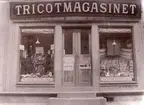 Tricotmagasinet, entrédörr och skyltfönster, skyltar med tyger och kläder.