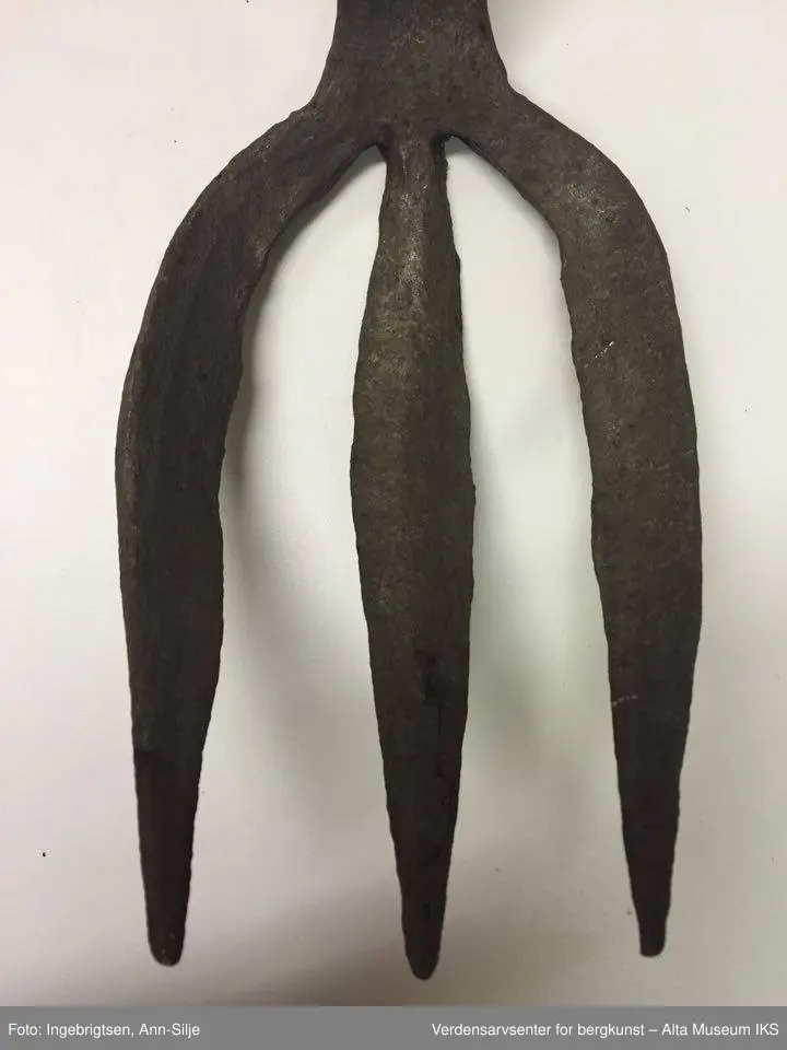 Trearmet jerngaffel. Kort skaft i samme metall. Liten krok på baksiden øverst på skaftet. To hull i skaftet (3 cm mellomtom). Hver av "armene" er ca. 20 cm lange. Skaftet er 23 cm langt.