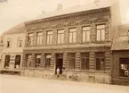 Regnellska huset vid Algatan 59 kvarteret Romulus Trelleborg, tomt nummer 4 (116) omkring år 1900, foto från N mot S. Längst till vänster i fönstret på andra våningen står ägaren häradsskrivaren L A Regnell och på trappan ser vi sonen Elvir, sedermera häradsskrivare i Örebro, 9149, huset vänster om är J: M. Möllers diverseaffär fastighet å tomt nummer 5 (114) och till höger byggmästare Joh. Holmgren å tomt nummer 3 (118A), nummer 26, 89:0901.