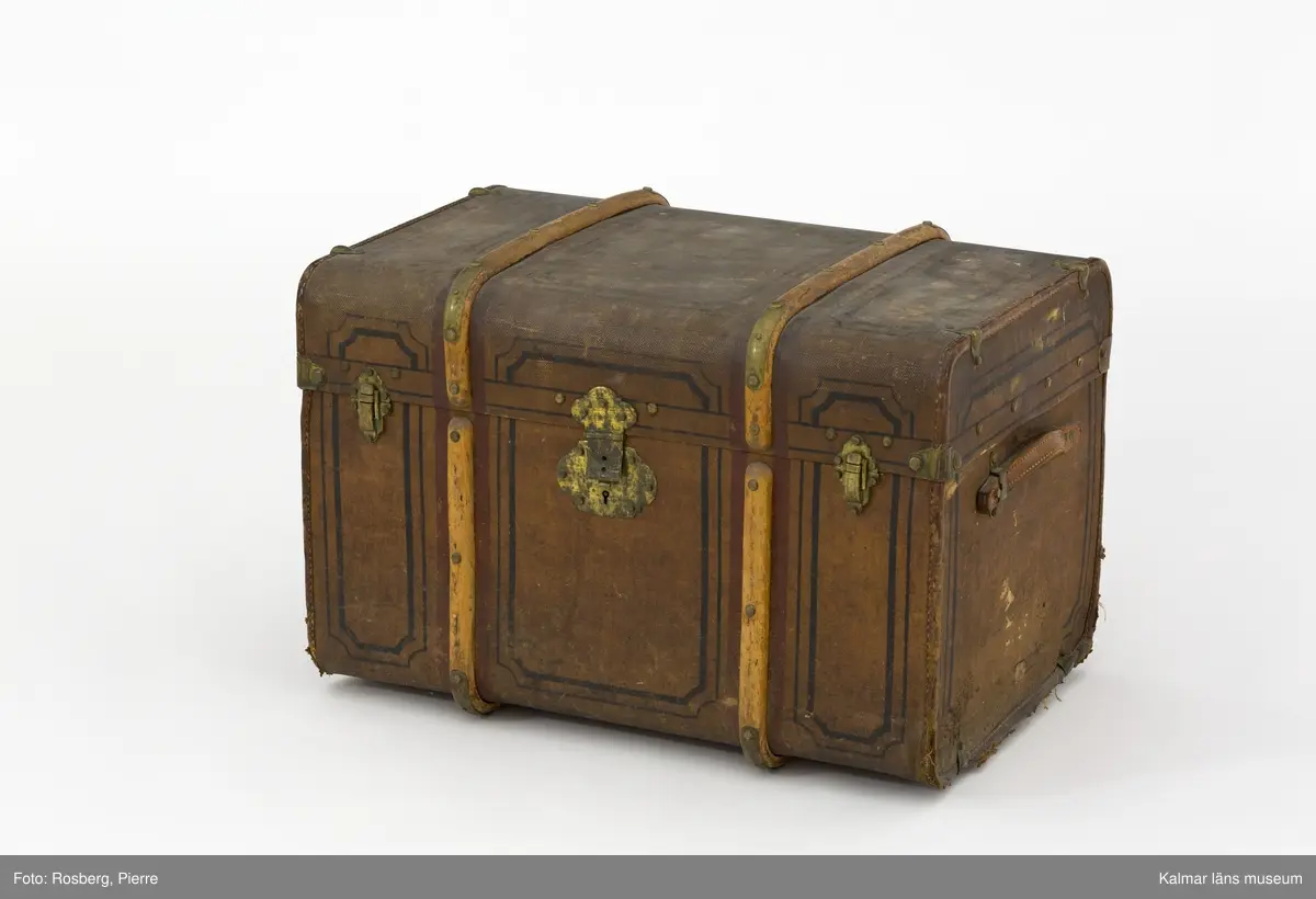 KLM 31996. Koffert, reskoffert, trä, metall, textil, läder, färg, Brunmålad, rektangulär koffert med svarta mönster och ett handtag av läder på varje gavel. På fronten ett nyckellås samt två låsspännen av mässing. Ribbor av trä med mässingsbeslag runt kofferten som skydd/stötdämpning. Inuti klädd med grön- och vitrandig papp med väv på baksidan.