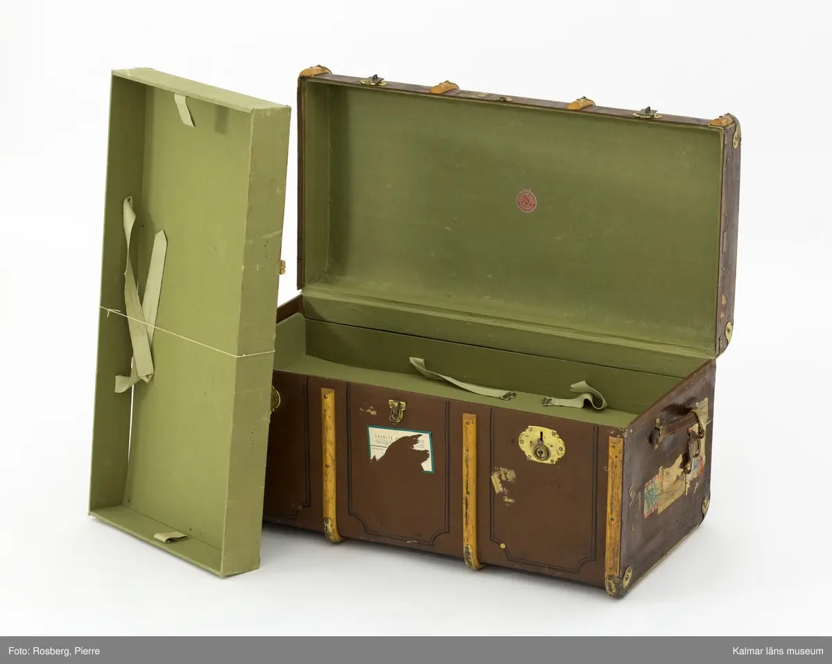 KLM 32819:4. Koffert, reskoffert, trä, metall, läder, textil. Brunmålad reskoffert av trä, belagd med väv, med svartmålad dekor. Beslag av trä och metall för stötdämpning/skydd. På varje gavel ett handtag av läder. På fronten tre låsspännen av mässing. Invändigt klädd i grönt tyg med två uttagbara förvaringskorgar av trä med spännen. I den ena är botten lös. På lockets insida klisterlapp: Senaste nyheter HANDSKAR PARAPLYER RESEFFEKTER SVEN NÄRSTAD KALMAR. 
Ovanpå locket etikett: PAKET-eller? Från Fru Hertha Jeansson, Kalmar, Kalmar C Efterkrav Kr..... Till Fru Märtha Frick Svedbergshuset Wennerbergsgatan 6 Bestämmelsestation STOCKHOLM (enligt fraktsedeln) 1 kolli.
Målat på lockets ovansida: M.J 3974 1.
På höger gavel tillverkarmärke: AKTIEBOLAGET JOHN A. GAHM GÖTEBORG,9.
