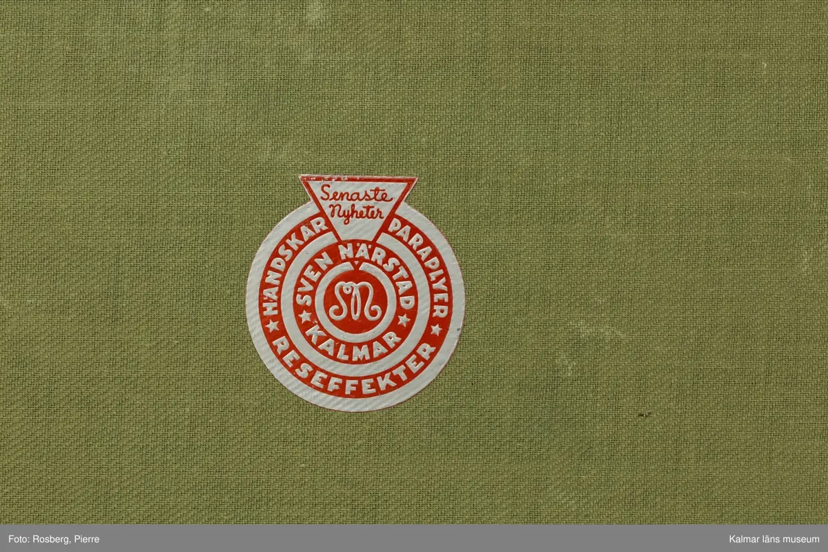 KLM 32819:4. Koffert, reskoffert, trä, metall, läder, textil. Brunmålad reskoffert av trä, belagd med väv, med svartmålad dekor. Beslag av trä och metall för stötdämpning/skydd. På varje gavel ett handtag av läder. På fronten tre låsspännen av mässing. Invändigt klädd i grönt tyg med två uttagbara förvaringskorgar av trä med spännen. I den ena är botten lös. På lockets insida klisterlapp: Senaste nyheter HANDSKAR PARAPLYER RESEFFEKTER SVEN NÄRSTAD KALMAR. 
Ovanpå locket etikett: PAKET-eller? Från Fru Hertha Jeansson, Kalmar, Kalmar C Efterkrav Kr..... Till Fru Märtha Frick Svedbergshuset Wennerbergsgatan 6 Bestämmelsestation STOCKHOLM (enligt fraktsedeln) 1 kolli.
Målat på lockets ovansida: M.J 3974 1.
På höger gavel tillverkarmärke: AKTIEBOLAGET JOHN A. GAHM GÖTEBORG,9.