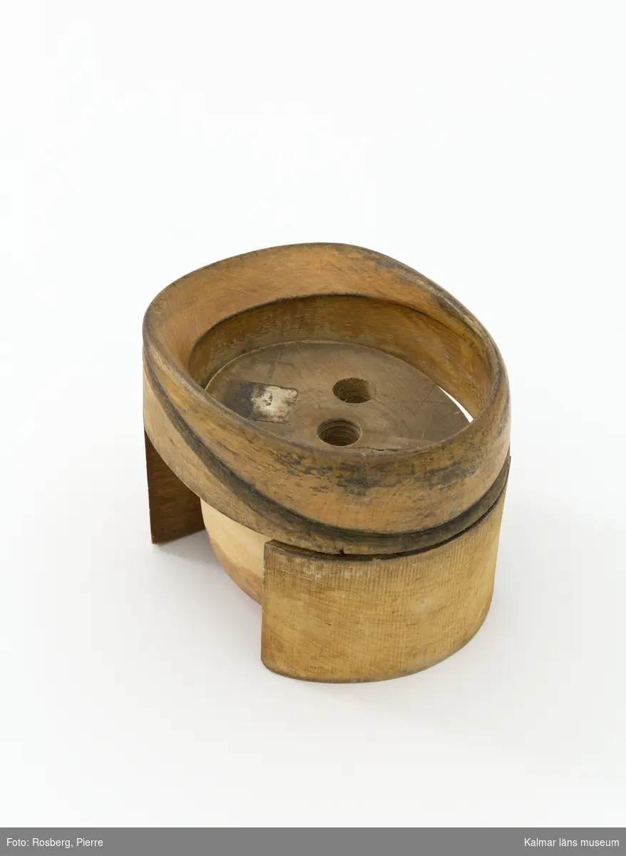 KLM 29821. Hattstock, damhattstock, herrhattsmodell. Hattstock av trä med uppvikt brätte baktill och sluttande brätte framtill. Veckad kulle som är något högre fram än bak. Hattstocken består av ett brätte på två fötter och en lös kulle som placeras i brättet. På kullens undersida är en lapp med oläslig text fastklistrad, texten har varit "En produkt från HATTSTOCKSFABRIKEN GÄLLSTAD". Bredvid finns även tre borrade hål med spår av textil som troligtvis har använts som handtag. Intill dessa finns kryss både inristat och skrivet med blyerts liksom siffran 807, liknande markeringar finns även på brättets kortsida. Kullen har slipats med sandpapper nedtill, så att ytbehandlingen försvunnit. Under hattillverkningen använde man sig av hattstockar för att få fram formen på hatten, det gjorde man genom att trä på stumpen eller kaplinen på stocken. Hattstockens form är därmed avgörande för hattens utseende. Denna modell användes vid tillverkningen av damhattar och är i form 807.