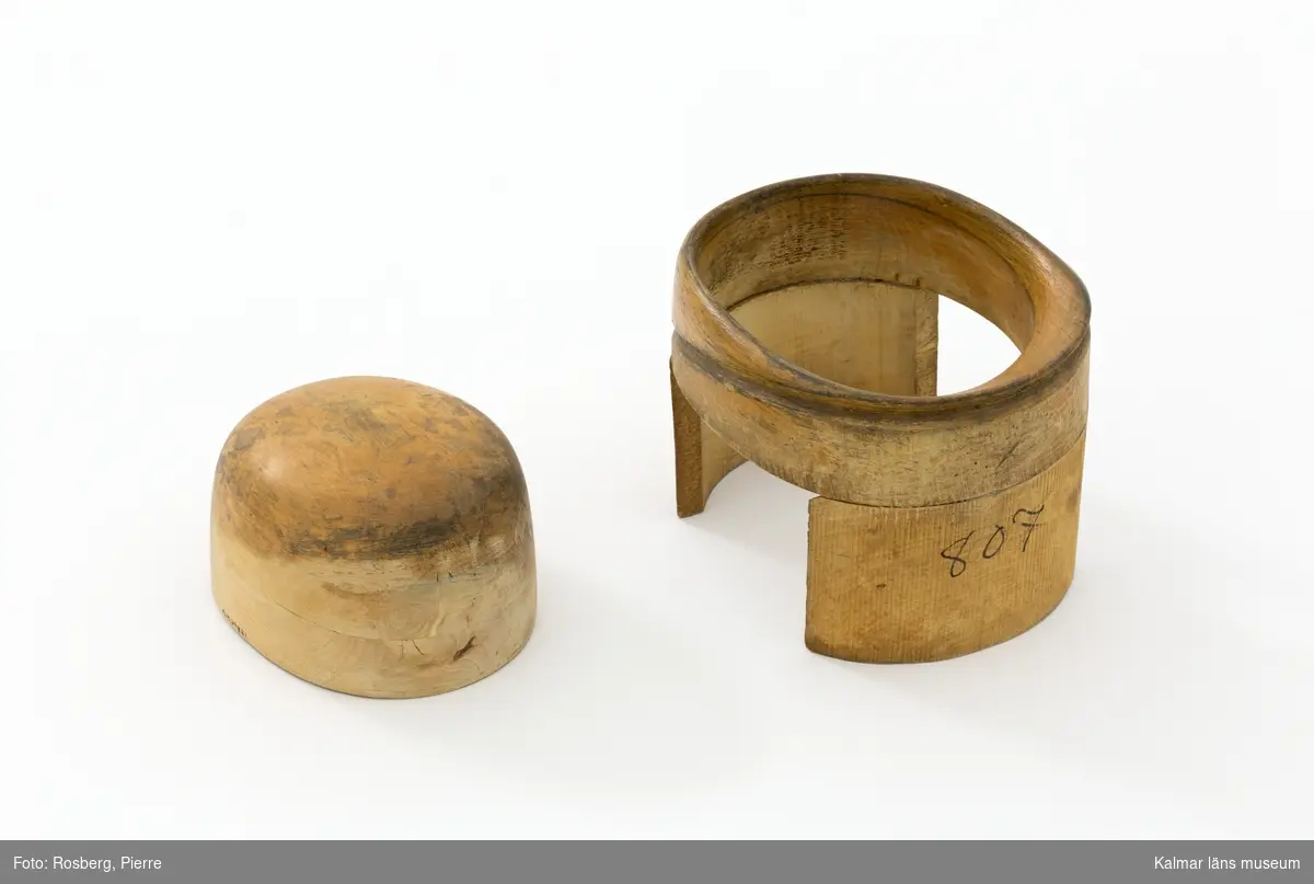 KLM 29821. Hattstock, damhattstock, herrhattsmodell. Hattstock av trä med uppvikt brätte baktill och sluttande brätte framtill. Veckad kulle som är något högre fram än bak. Hattstocken består av ett brätte på två fötter och en lös kulle som placeras i brättet. På kullens undersida är en lapp med oläslig text fastklistrad, texten har varit "En produkt från HATTSTOCKSFABRIKEN GÄLLSTAD". Bredvid finns även tre borrade hål med spår av textil som troligtvis har använts som handtag. Intill dessa finns kryss både inristat och skrivet med blyerts liksom siffran 807, liknande markeringar finns även på brättets kortsida. Kullen har slipats med sandpapper nedtill, så att ytbehandlingen försvunnit. Under hattillverkningen använde man sig av hattstockar för att få fram formen på hatten, det gjorde man genom att trä på stumpen eller kaplinen på stocken. Hattstockens form är därmed avgörande för hattens utseende. Denna modell användes vid tillverkningen av damhattar och är i form 807.