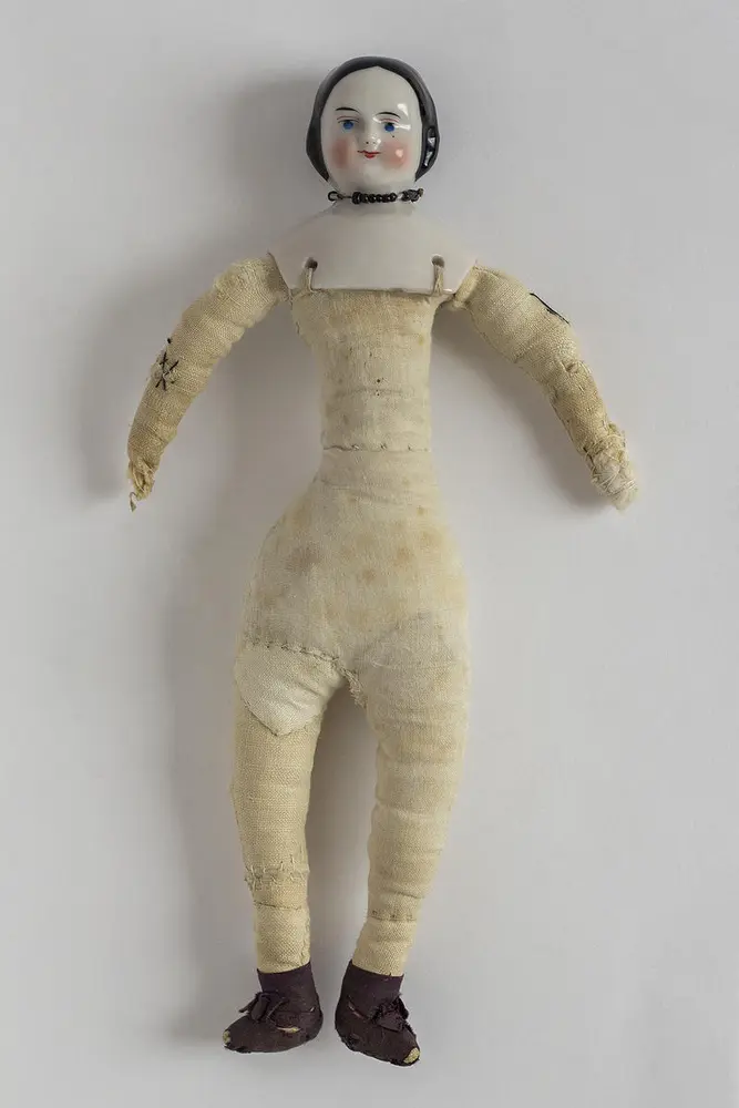 Den hjemmelagede dukkekroppen er sydd sammen av mange stoffbiter av ulike lerretskvaliteter og stoppet med et mjukt materiale, og de slitte dukkehendene er reparert med hvit sytråd.