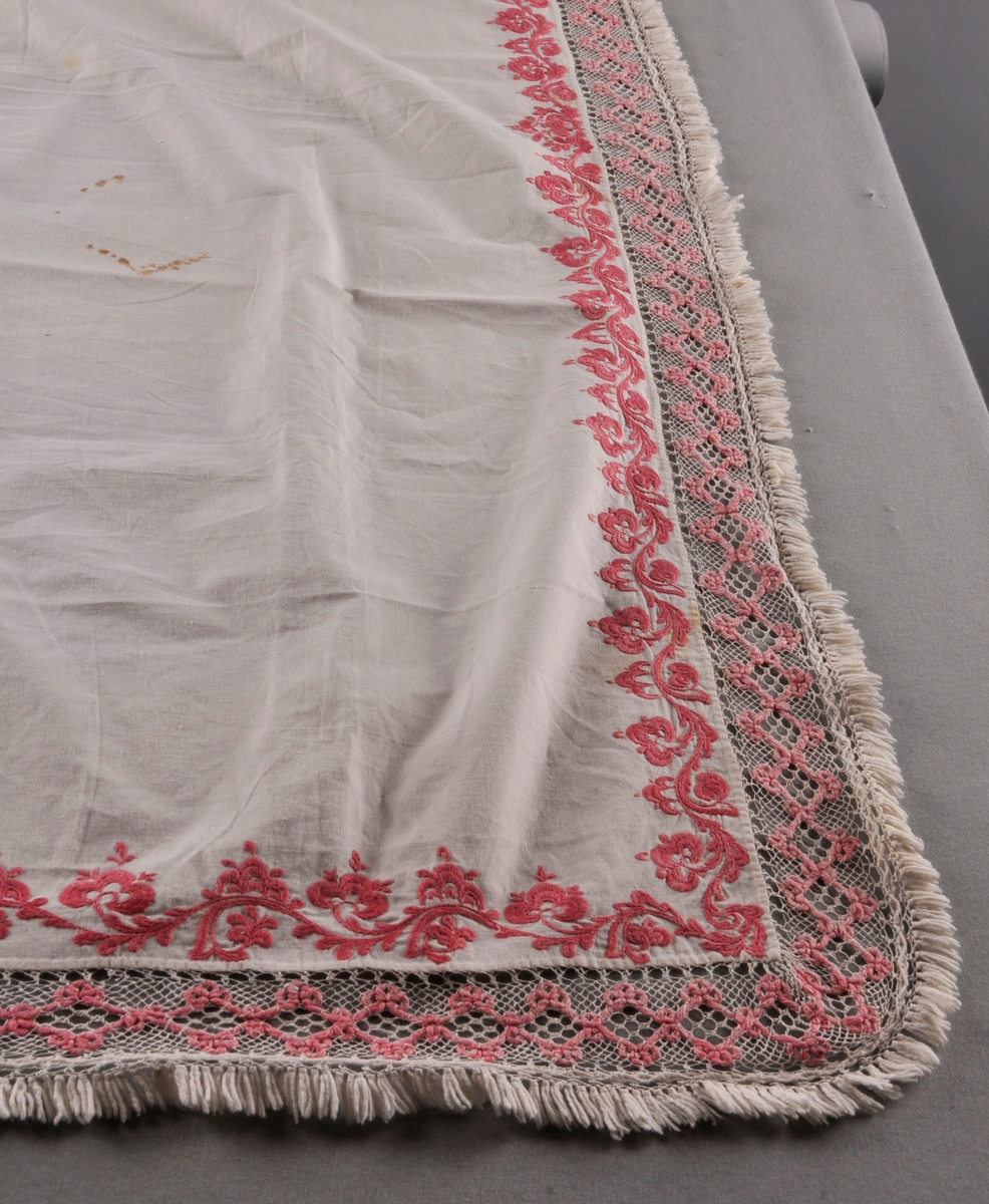 Rektangulært stykke av bomullslerret med rosa rosesaum kring alle sider. Ei påsauma kvit og rosa blonde i Telemarksbinding, i to ulike bredder, langs alle sidene.