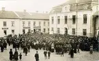 Livsmedelsdemonstration i Falköping inför stadsfullmäktige. På trappan står stadsfullmäktiges ordförande rektor Hagberg, borgmästare Koch, veterinär Kylén och disponent Victor Karlson. Talare är hr Ernst Sandell och framför honom står handl. Fredrik Westlin. Gåva till Falköpings Stadsarkiv.