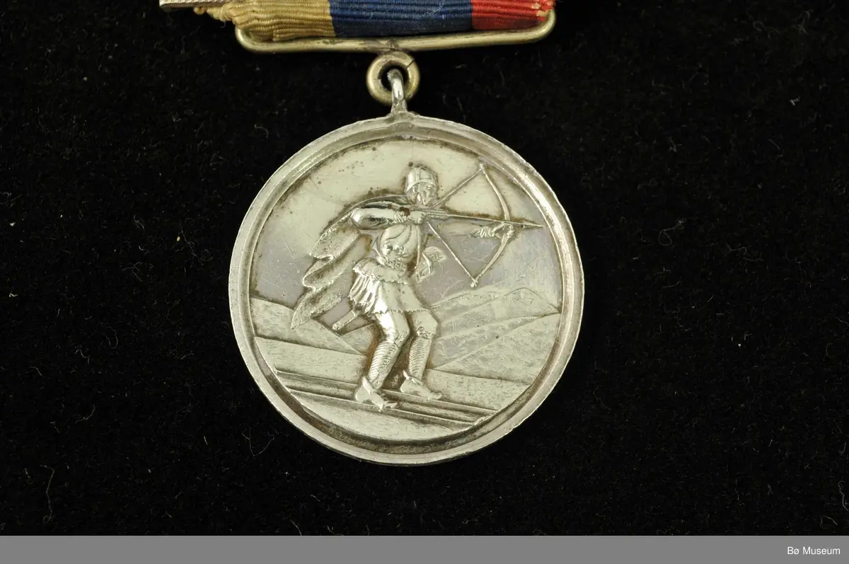 Rund medalje med bånd og nål. Skiløper med pil og spent bue på den ene siden, på den andre tekst.
