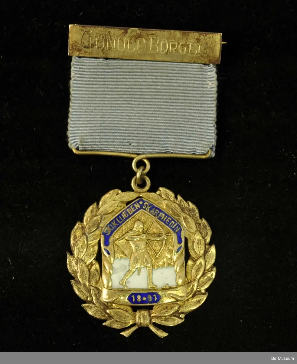 Medalje med lyseblått bånd, navneplate og laurbærkrans rundt et merke med skigåer med pil og spent bue.