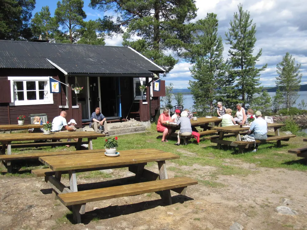 Oversiktsbildet av sletta foran huset museet bruker som kafé- og resepsjonslokale. Det sitter personer spredt rundt på bordene på sletta.