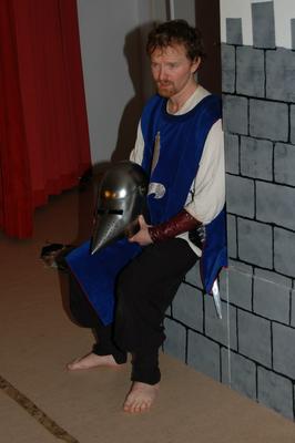 Mann i middelalderklar som holder en metallhjelm. Svarte bukser, blå surcoat, hvit skjorte og lær armbeskyttere