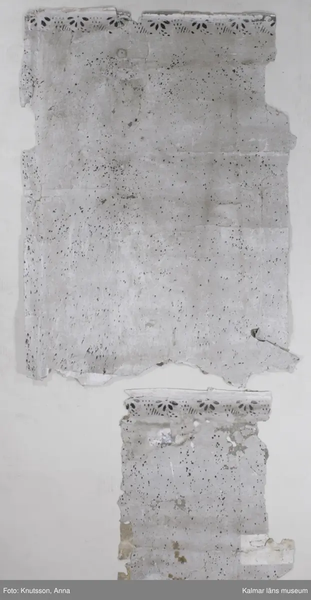 KLM 43871 Tapet i papper. 13 bitar. Mått: minsta 8 x 4 cm. Ljusgrå stänktapet med stänk i svart, med schablonmålad bård i svart på samma grå bakgrund. Bitarnas baksida har motiv från tre olika tapeter. En är målad med stora blommönster i dova färger: rosa, vitt, grönt och rött med blommor i lysande cinnoberrött. En annan har mustigt gröna bladrankor i diagonalrutor, med mustigt gröna blommor inuti. Tapeten har grå botten. Den tredje har vit botten med spridda blomsterbuketter i klara färger, gröna blad med blommor i rött och orange-gult. Datering: De målade tapeterna är 1700-tal. Någon gång under 1800-talet har man vänt på tapeterna och stänkmålat dem.