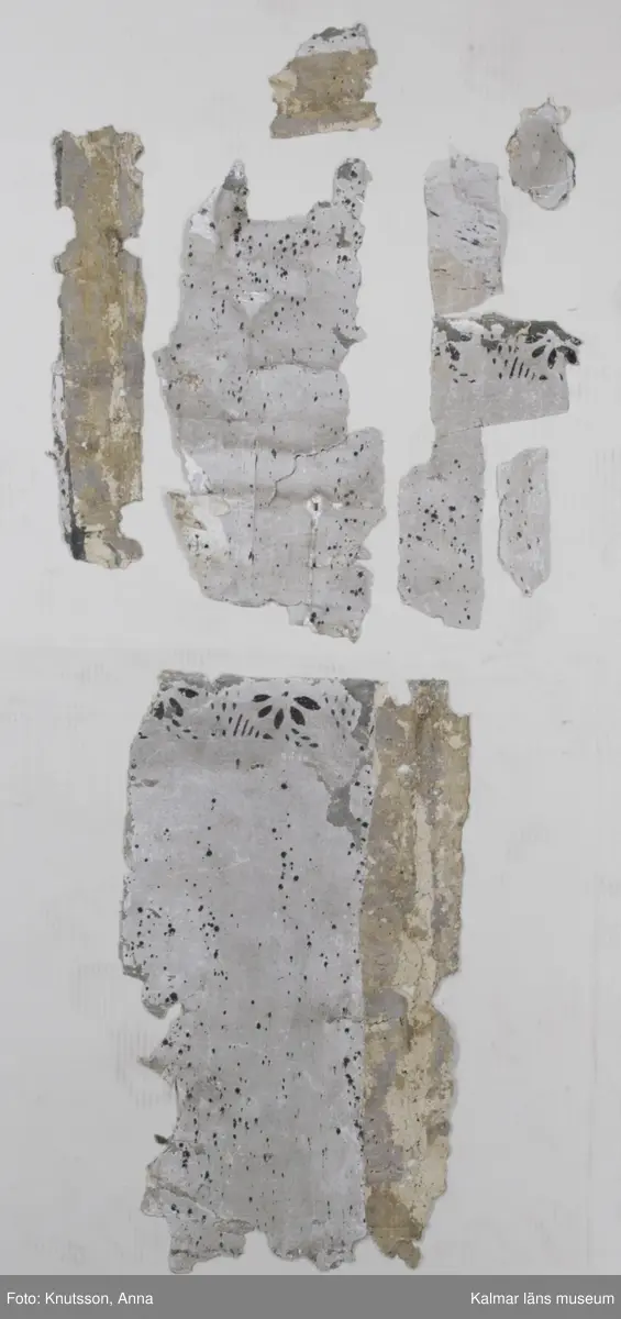 KLM 43871 Tapet i papper. 13 bitar. Mått: minsta 8 x 4 cm. Ljusgrå stänktapet med stänk i svart, med schablonmålad bård i svart på samma grå bakgrund. Bitarnas baksida har motiv från tre olika tapeter. En är målad med stora blommönster i dova färger: rosa, vitt, grönt och rött med blommor i lysande cinnoberrött. En annan har mustigt gröna bladrankor i diagonalrutor, med mustigt gröna blommor inuti. Tapeten har grå botten. Den tredje har vit botten med spridda blomsterbuketter i klara färger, gröna blad med blommor i rött och orange-gult. Datering: De målade tapeterna är 1700-tal. Någon gång under 1800-talet har man vänt på tapeterna och stänkmålat dem.