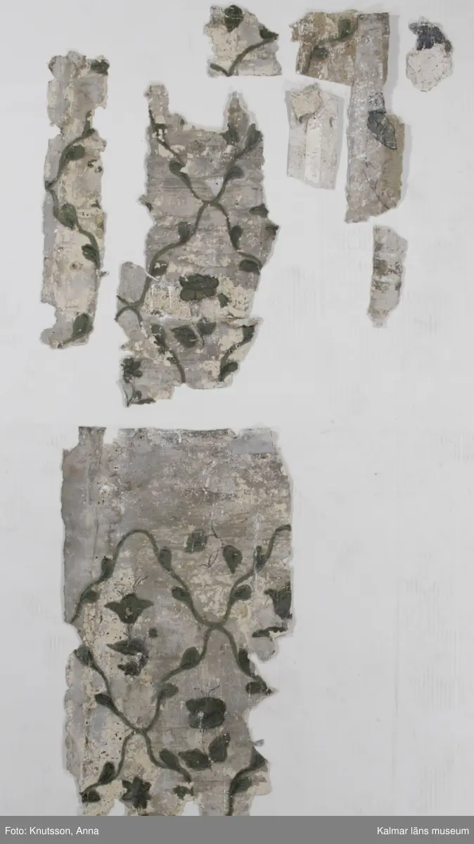KLM 43871 Tapet i papper. 13 bitar. Mått: minsta 8 x 4 cm. Ljusgrå stänktapet med stänk i svart, med schablonmålad bård i svart på samma grå bakgrund. Bitarnas baksida har motiv från tre olika tapeter. En är målad med stora blommönster i dova färger: rosa, vitt, grönt och rött med blommor i lysande cinnoberrött. En annan har mustigt gröna bladrankor i diagonalrutor, med mustigt gröna blommor inuti. Tapeten har grå botten. Den tredje har vit botten med spridda blomsterbuketter i klara färger, gröna blad med blommor i rött och orange-gult. Datering: De målade tapeterna är 1700-tal. Någon gång under 1800-talet har man vänt på tapeterna och stänkmålat dem.