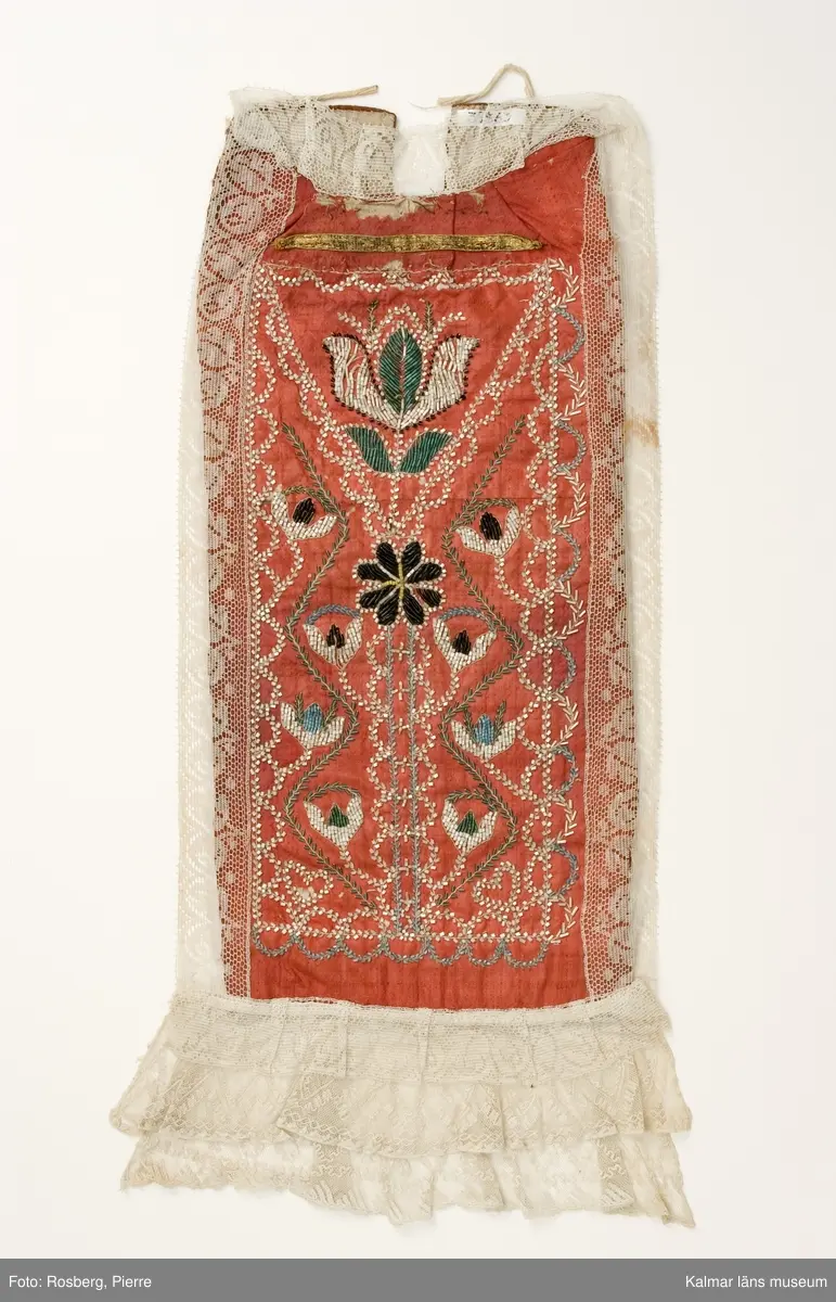 KLM 39363. Doppåse. 1800-tal. Sydd av rött, glättat ylletyg s.k. rask. Hela framsidan är broderad med små vita, svarta, gröna och ljus blå glaspärlor. Blommönster och girlanger. Ett smalt band av gulmetall är fastsytt ovanför pärlbroderierna, nära halsringningen. Halsringning, långsidor och nederkant är kantade med vit maskintillverkad spets. Vid nederkanten är två lager av vit broderad tyllspets fastsydd. Bakstycke av rödbrunt, tuskaftat ylletyg. Ett ca 19 cm långt sprund, från halshålet och ner, mitt bak. Knytband av linne fastsydda vid överkanten. Fodrad med vitt tuskaftat linne.