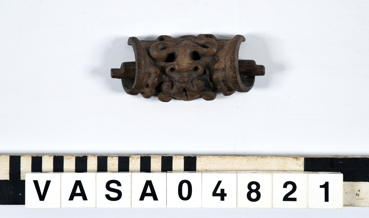 Rullverksornament.
Skulpterat litet, spänneliknande ornament, dekorerat i broskverksstil.
Det långsmala ornamentet är format som ett stycke kraftigt upprullad bit näver eller läder, med konkav bak- eller undersida. På baksidan är ornamentet profilerat för en list. Broskdekoren på ovansidan är formad som en maskaron.
Ornamentet är mycket välbevarat.

Text in English: A small, buckle-like ornament, decorated in a style similar to earshell work.
The long narrow ornament is formed as a piece of tightly rolled bark or leather, with a concave back or underside so as to match a batten or the like. The reverse side is profiled to fit a moulding. The earshell work on the upper side is shaped as a mask.
The ornament is well preserved.