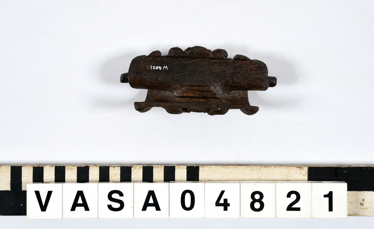 Rullverksornament.
Skulpterat litet, spänneliknande ornament, dekorerat i broskverksstil.
Det långsmala ornamentet är format som ett stycke kraftigt upprullad bit näver eller läder, med konkav bak- eller undersida. På baksidan är ornamentet profilerat för en list. Broskdekoren på ovansidan är formad som en maskaron.
Ornamentet är mycket välbevarat.

Text in English: A small, buckle-like ornament, decorated in a style similar to earshell work.
The long narrow ornament is formed as a piece of tightly rolled bark or leather, with a concave back or underside so as to match a batten or the like. The reverse side is profiled to fit a moulding. The earshell work on the upper side is shaped as a mask.
The ornament is well preserved.