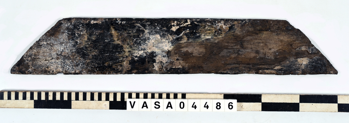 Odekorerad trapetsoid med bort-vittrade tappar på båda ändarna.  Fyra spikhål. En ände devlis avsplittrad. Mycket vittrad och torr yta. Tjockleken på brädan vid tredje spiken från vänster väsentligt mycket tjockare än resten, möjligtvis originaltjocklek.