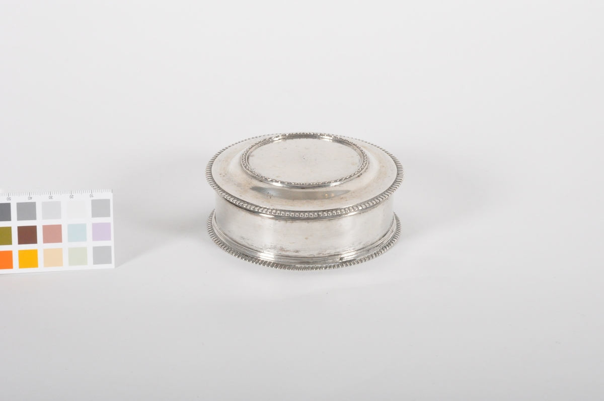Sirkulær obalteske i sølv med avtagbart lokk. Dekorert med liten bård i ytterkant av lokk og langs bunn på eske. Tynn, forhøyet sik-sakbård oppå lokket.