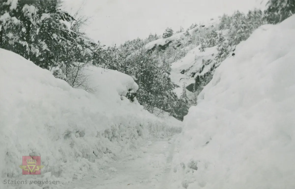 Bildet er fra hovedveiene rundt i Vest-Agder vinteren 1937. Store snømengder. Ref. til "Meddelelser fra Veidirektøren", Nr. 3-1937. "De vanskelige sneforhold på Sørlandet i år". Av avdelingsingeniør T. Backer. Se vedlegg i Nedlastinger. 

31.03.2017: "Bildet er tatt 200 meter ovenfor fyllinga på Utsikten i Kvinesdal på vei østover og plassen heter Skohauen, det var her kirkefolket skiftet over til fine sko og gikk ut av treskoene. "

"Det andre bilde som er samlet sammen med bilde overfor nevnt, er tatt på samme sted ved Skohauen overfor Utsikten i Kvinesdal, og viser 2 personer og bil, fotograf Colbjørnsen har møtt en bil på smal vei. Vinteren 1937-38."