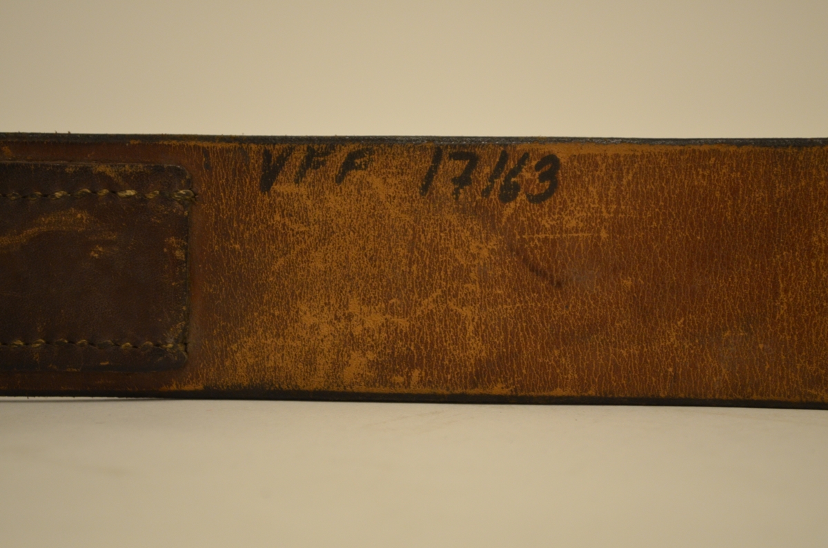 Brunsvart belte tilhørende uniform brukt av den tyske okkupasjonsmakten i Norge under andre verdenskrig. Beltet er av lær med beltespenne i metall. Emblemet med den tyske ørn og hakekors er skrapet bort fra beltespennen. Stempel på innsiden av læret, hvor "Aachen 1942" kan tydes. Varemerke utydelig.