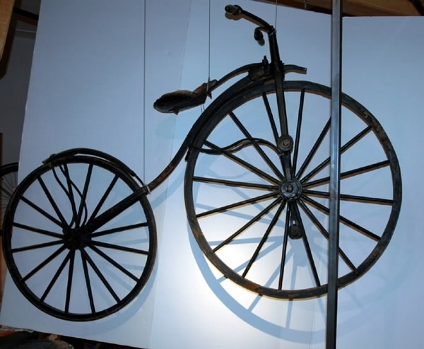 Cykel, Michaux-typ av järnsmide. Avancerad typ. Svagt S-formig ram med högt uppdraget styrrör och skålformig sits av trä, på separat fjäderblad. Bakåtsvept styrstång med åt sidorna rakt utstående handtag av furu. Stänkskärmar. Järnskodda hjul av trä på dubbel rad ekrar. Trampor av trä, på järnaxlar, justerbart fastskruvade i vevar, direkt verkande på framaxeln. Svart med röd sits.