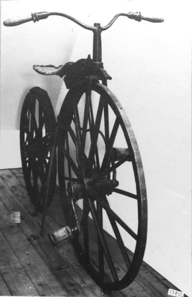 Cykel, Michaux-typ av järnsmide. Avancerad typ. Svagt S-formig ram med högt uppdraget styrrör och skålformig sits av trä, på separat fjäderblad. Bakåtsvept styrstång med åt sidorna rakt utstående handtag av furu. Stänkskärmar. Järnskodda hjul av trä på dubbel rad ekrar. Trampor av trä, på järnaxlar, justerbart fastskruvade i vevar, direkt verkande på framaxeln. Svart med röd sits.