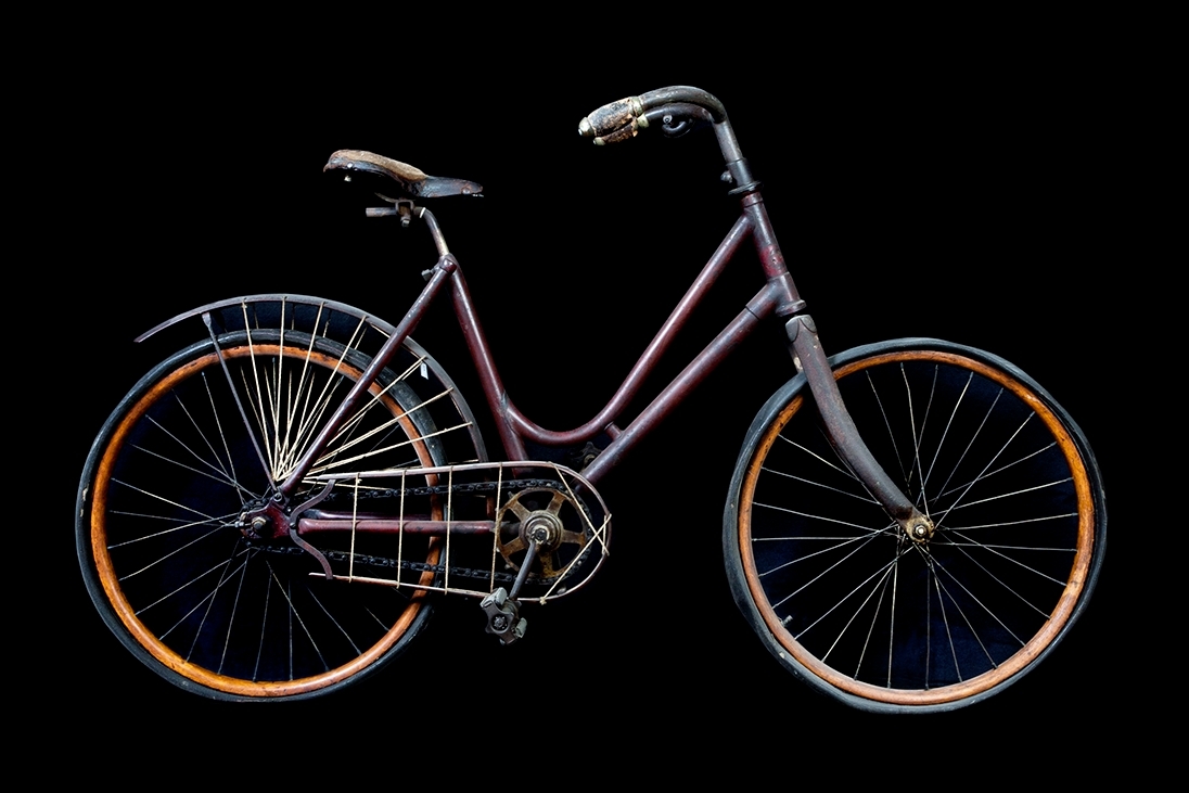 Cykel, flickstorlek av stålrör. Bakskärm och fälgar av trä. Stoppad lädersadel. Styre med korkhandtag och ringklocka. På bakskärmen hål för hjulskydd. Kedjeskydd av trä med hål för nät eller dylikt. Kedja av 2,4 cm långa länksidor, förenade genom massiva mellanstycken. Pedaler av gummi. Luftfyllda gummidäck. Ekrarna sammanlödda två och två vid navet. På styrrörets framsida firmamärke, se bild. Cykeln saknar broms och frihjul.