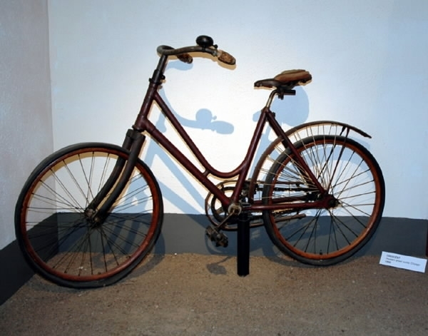 Cykel, flickstorlek av stålrör. Bakskärm och fälgar av trä. Stoppad lädersadel. Styre med korkhandtag och ringklocka. På bakskärmen hål för hjulskydd. Kedjeskydd av trä med hål för nät eller dylikt. Kedja av 2,4 cm långa länksidor, förenade genom massiva mellanstycken. Pedaler av gummi. Luftfyllda gummidäck. Ekrarna sammanlödda två och två vid navet. På styrrörets framsida firmamärke, se bild. Cykeln saknar broms och frihjul.