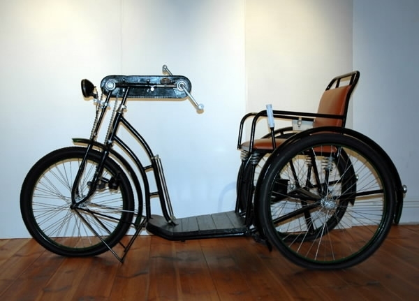 Trehjulscykel, rullstolscykel, svart med röd/brun sits i skinnimitation. Tre hjul som drivs med handvev genom en kedja till framhjulet. Handbroms.  Reglerbart ryggstöd. Fotplatta av trä, svart. Vit börs eller väska på sidan av sitsen.