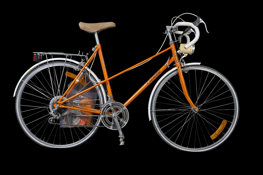 Crescent-cykel med racerstyre från Monark-Crescent AB i Varberg a) Orangefärgad 12-växlad damcykel med racerstyre/bockstyre. Däcken är ej original. Märkning: "306 Fashionline" Sadel: "Selle Royal".
b) Sadelskydd i skinn, 25x13 cm. c) Sadelskydd i orangefärgad plast, 24x13 cm. d) Röd/gul packrem med vita plasthakar, längd 52 cm. e) Ventil, metall, längd 2,5 cm.
f) Glasögon med vita skalmar och orangefärgade bågar, 50x130 mm, 1980-tal.