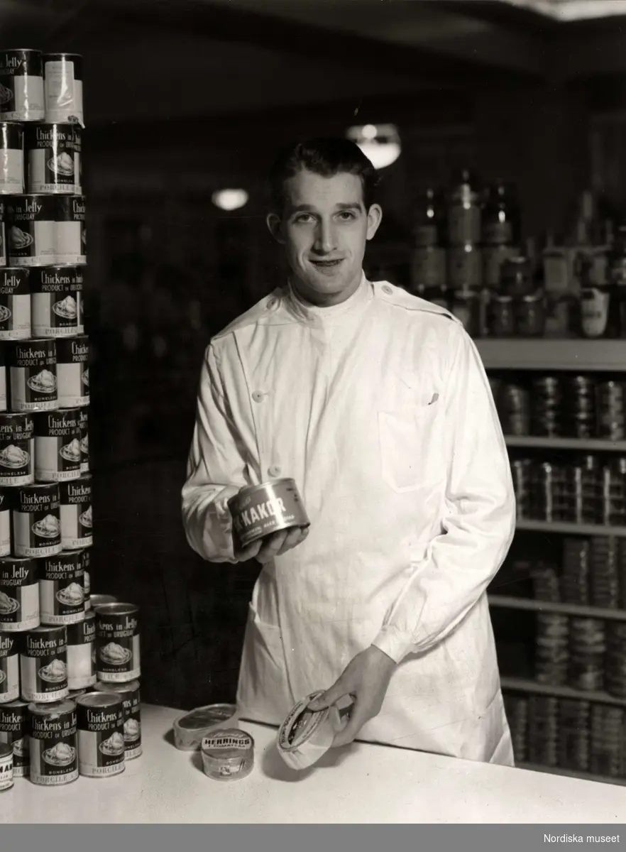 Manlig expedit i den nya arbetsrocken, livsmedelsavdelningen på varuhuset Nordiska Kompaniet i Stockholm 1947. Den leende unge mannen håller fram en burk, sannolikt innehållande fisk- kakor, framför honom på bordet ligger fler konservburkar med bland annat makrill. Vid hans sida har någon byggt en pyramid av konservburkar med Chicken in jelly, kyckling i gelé. I bakgrunden hyllor där burkar står uppradade.