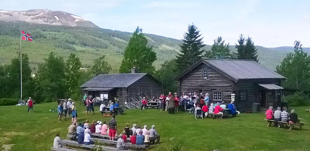 Oversiktsbilde av tunet med Buflodstua og Målstua. I bakgrunnen er Trysilfjellet. På tunet er det flere personer som står i ulike grupper.